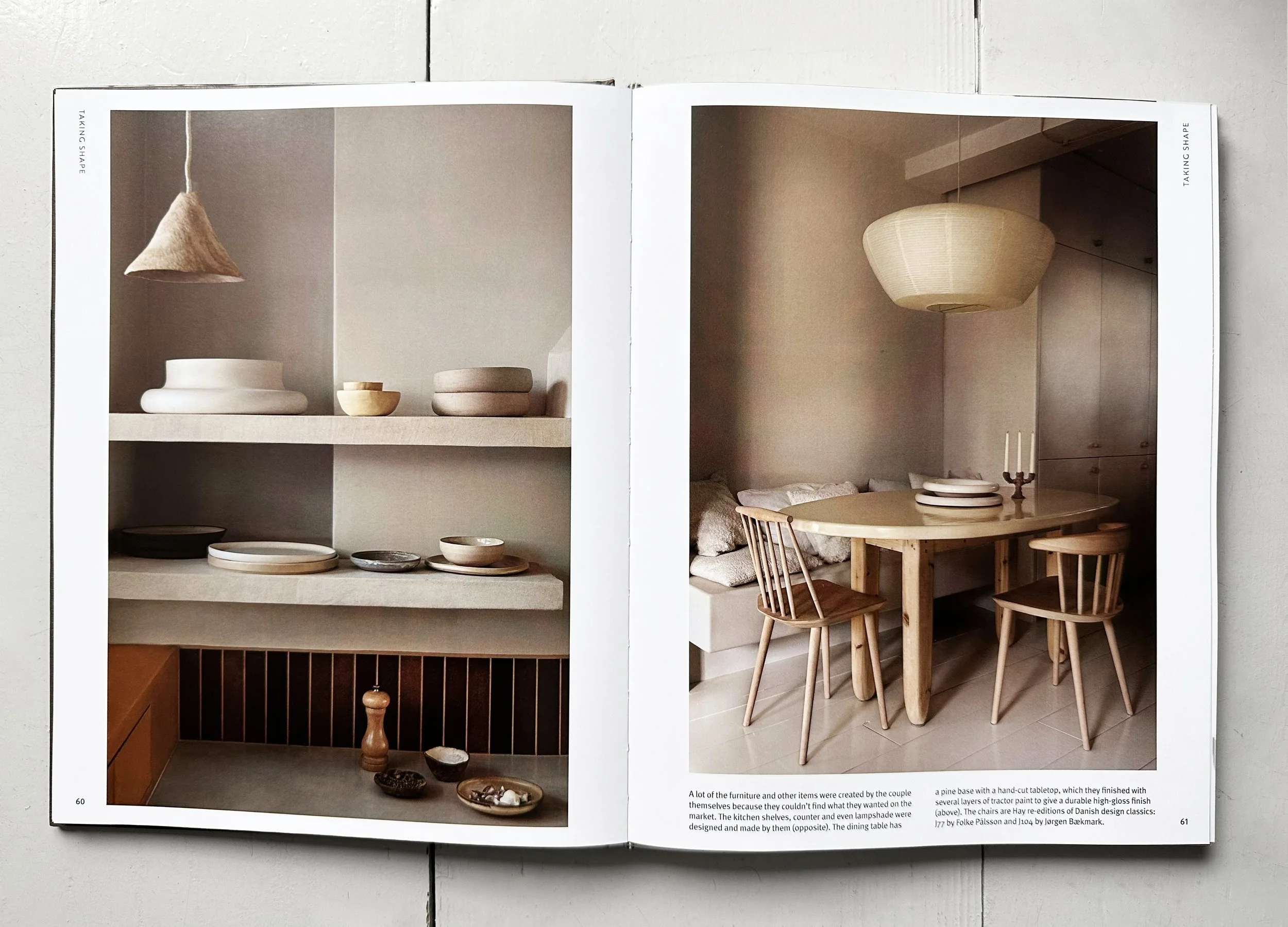 Open book displaying images of modern, minimalist interior design with neutral tones, featuring shelves with ceramics and a cozy dining nook with a round wooden table and chairs, and hanging lamps.