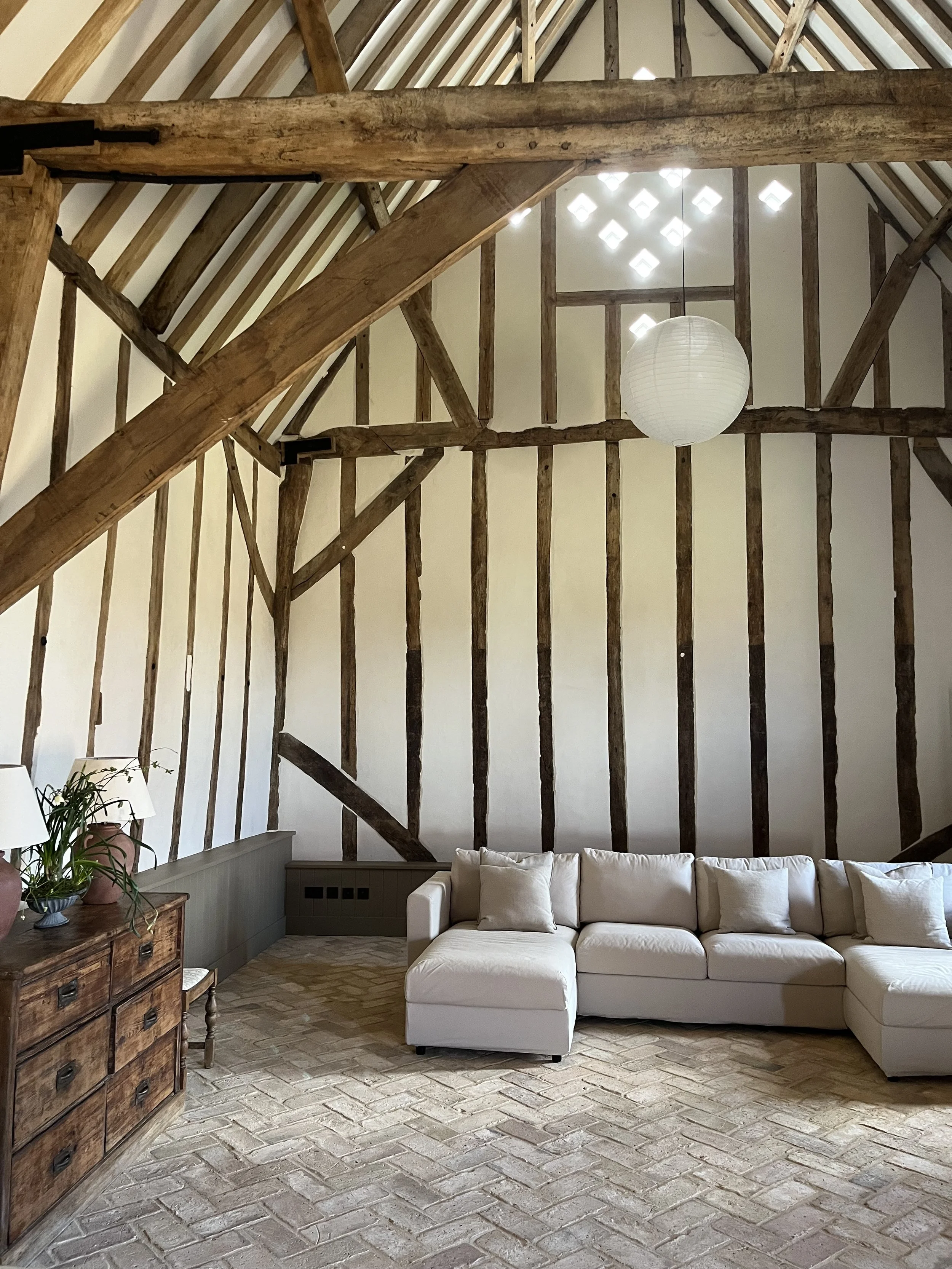 Living room with exposed wooden beams, white sofa with beige pillows, wooden side table with potted plant, brick floor, and paper lantern ceiling light.