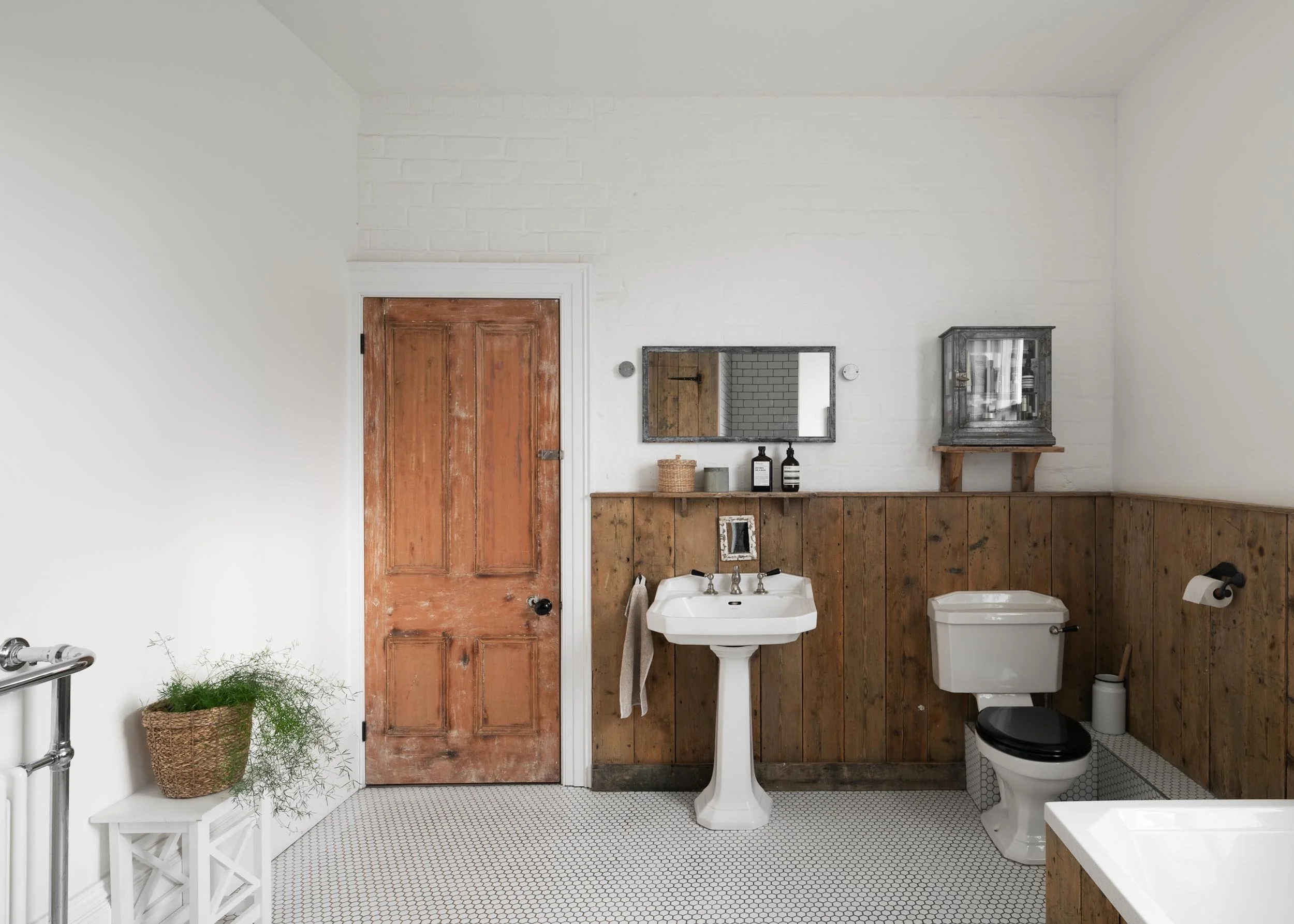 Bathroom with white walls, wooden door, pedestal sink, toilet with black seat, mirror, small shelves, decorative items, and a potted plant.
