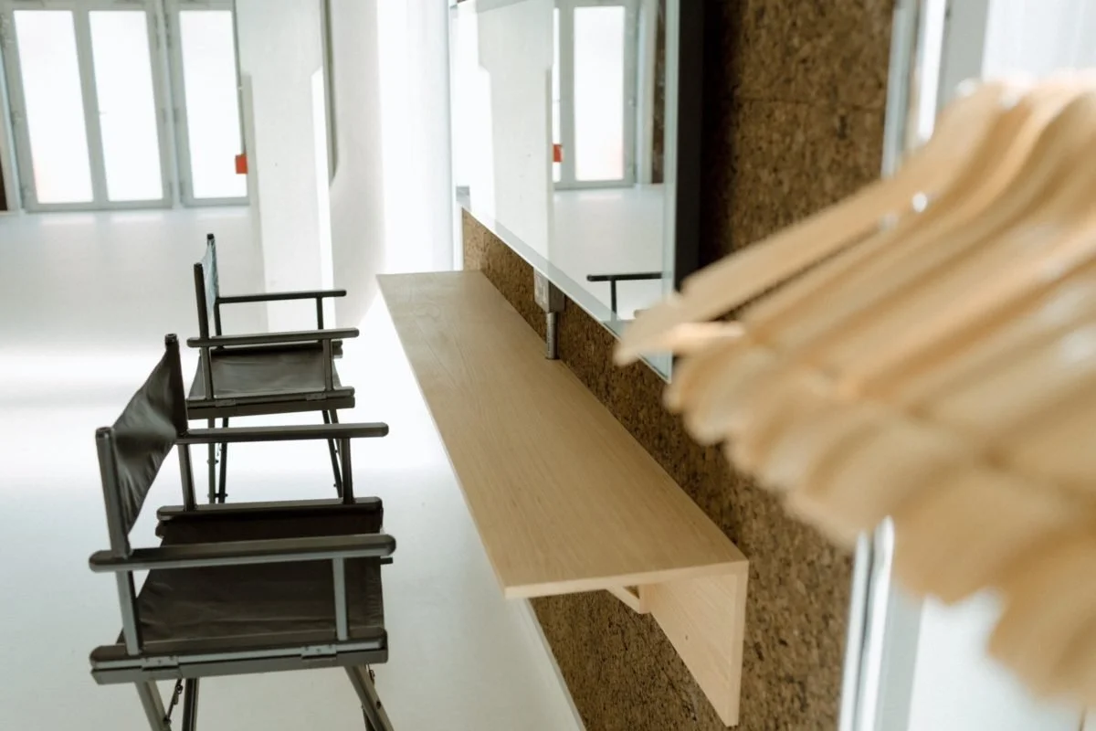 Interior view of a modern room with a long wooden desk, two black chairs, and a large mirror on the wall. Large windows let in natural light.