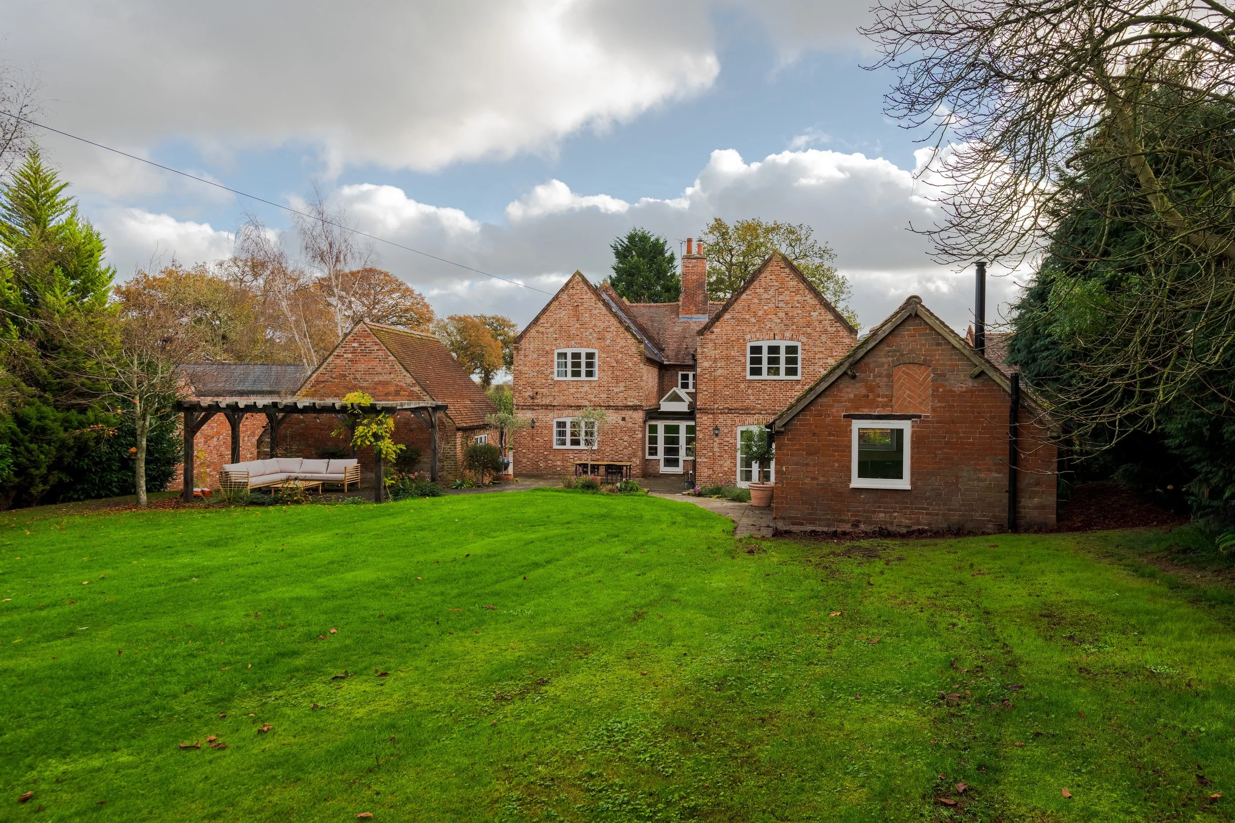 A brick house with a lush green lawn in the foreground and a garden swing under a wooden pergola on the left side, surrounded by trees and a cloudy sky.