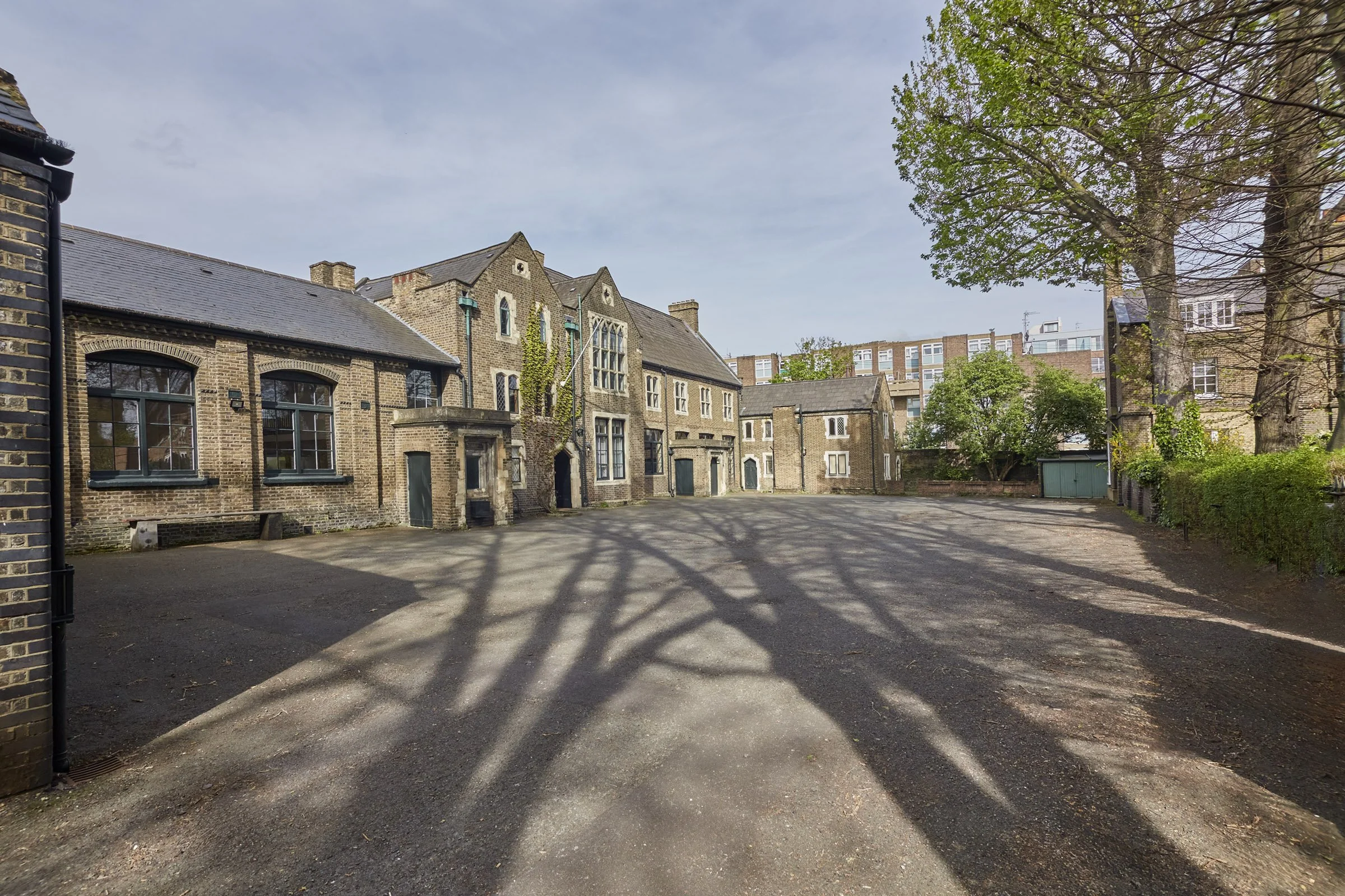 An empty open lot with shadows of tree branches on the ground, surrounded by historic brick buildings with diverse window styles and greenery, under a partly cloudy sky.