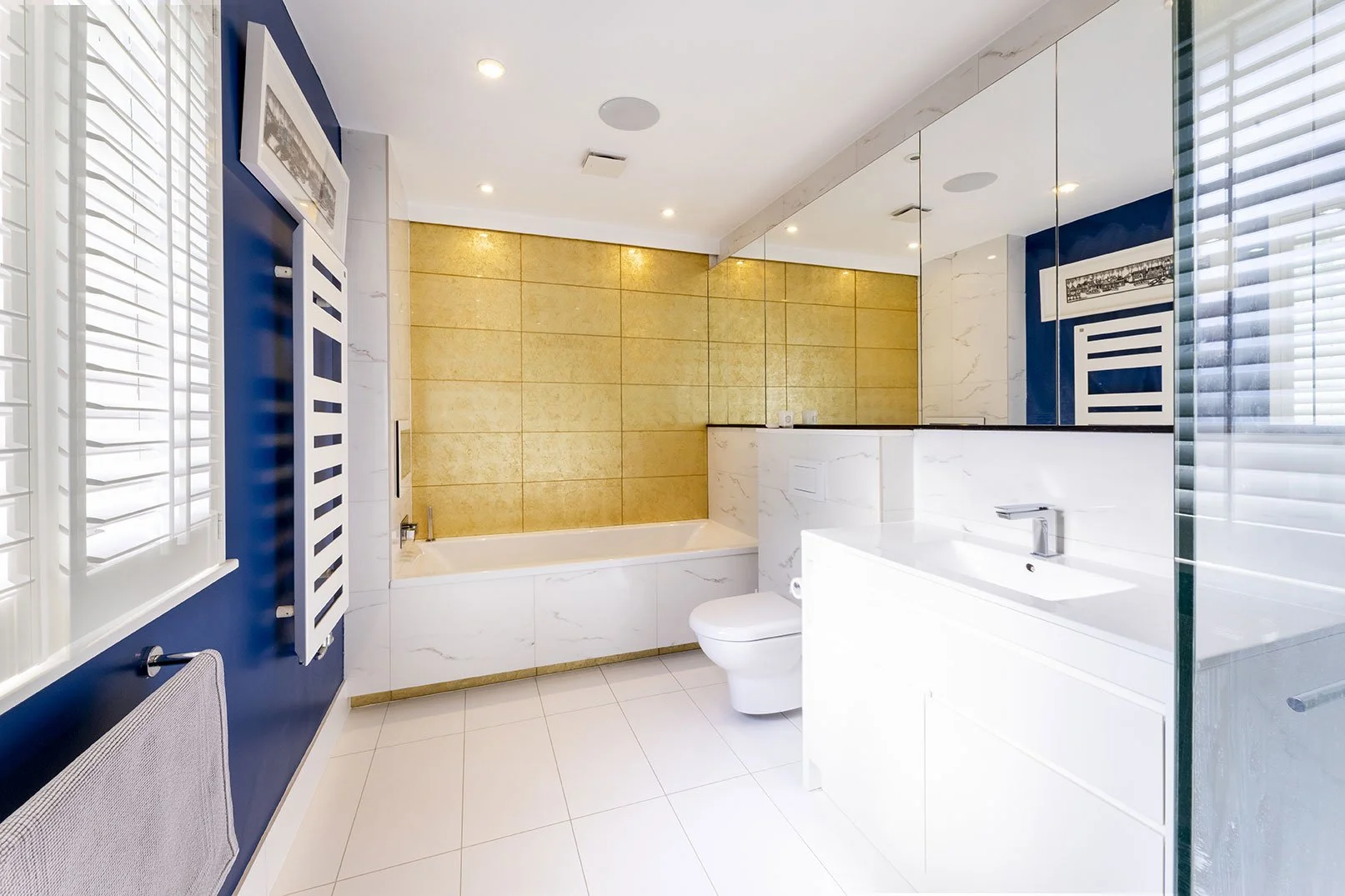 Modern bathroom with white marble finishes, a bathtub against a gold accent wall, a white toilet, and a white vanity with a sink. Blue walls with white window shutters and framed artwork.