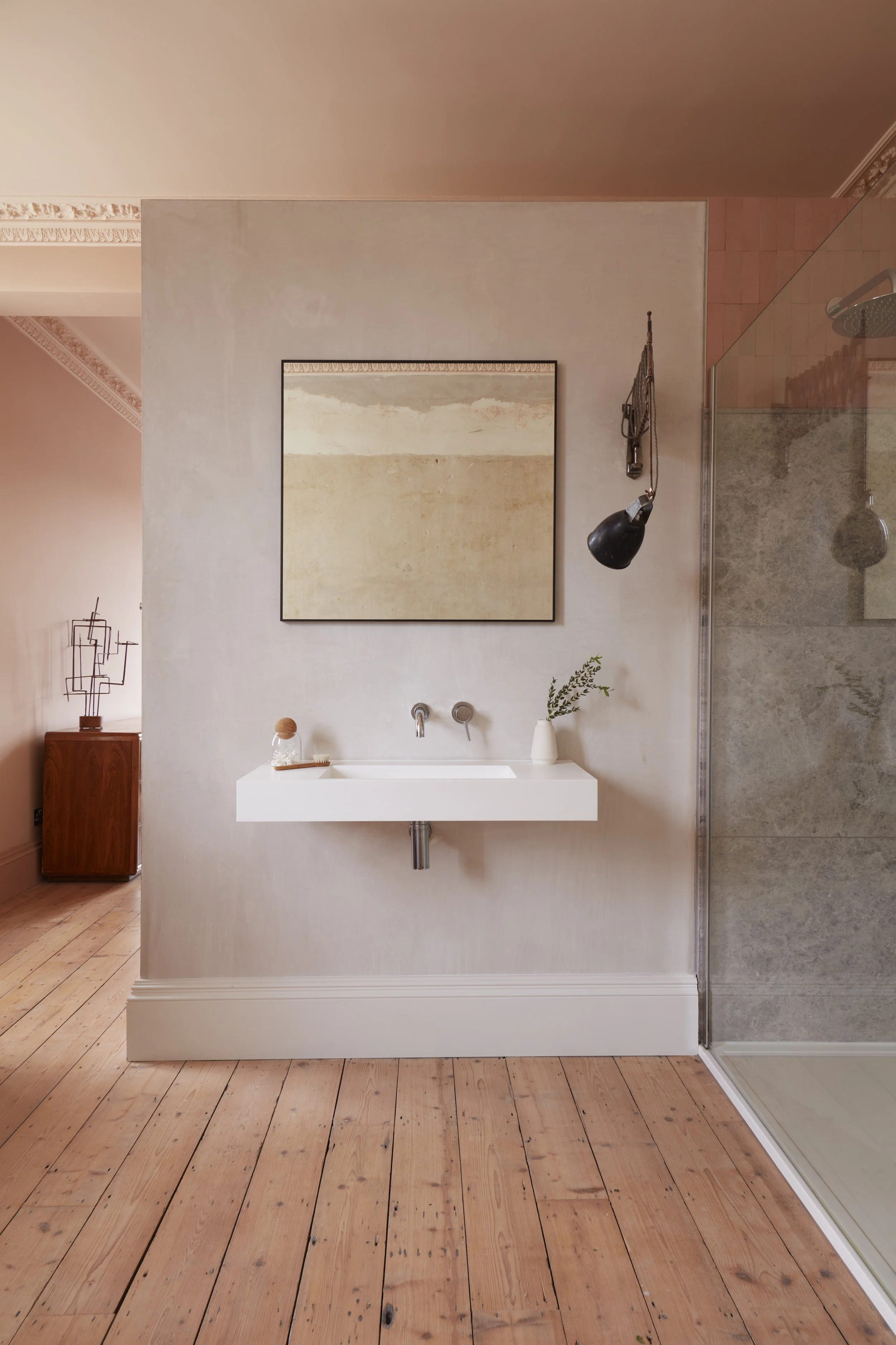 Minimalist bathroom with a floating white sink, a large beige abstract painting, a black hanging pendant lamp, a small white vase with greenery, and a corner shower with gray tiles on the right. Wooden flooring and light-colored walls complete the sc