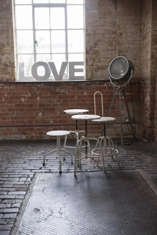 A cluster of white metal stools and a table on a tiled floor in an industrial-style room with exposed brick walls and a large window. A lamp stands beside the window with the word 'LOVE' displayed on the window.