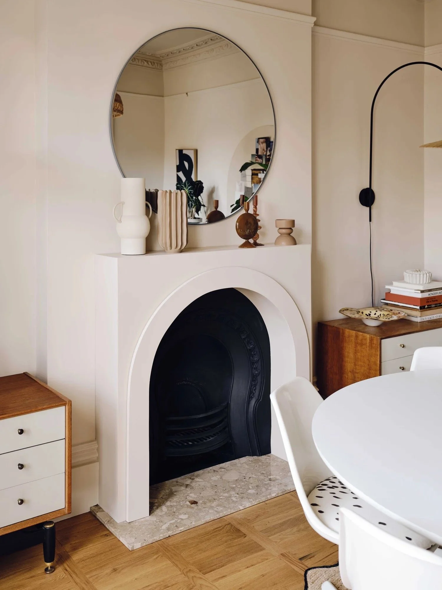 Living room corner with a white fireplace, decorated with vases and sculptures on the mantel and a large round mirror above it. There is a wooden cabinet with books and a decorative dish on the right side of the fireplace, and a white dining table wi