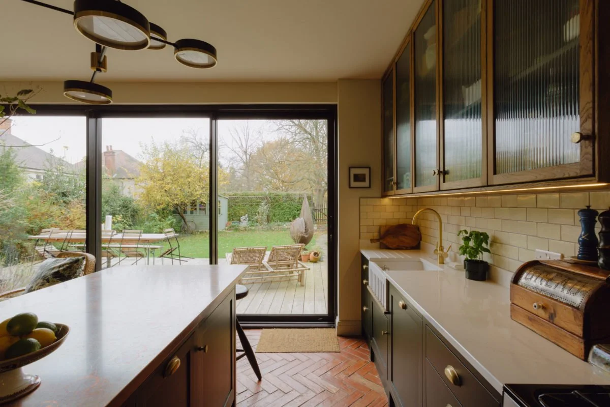 A modern kitchen with a large island counter and sliding glass door leading to a backyard with outdoor furniture and trees.