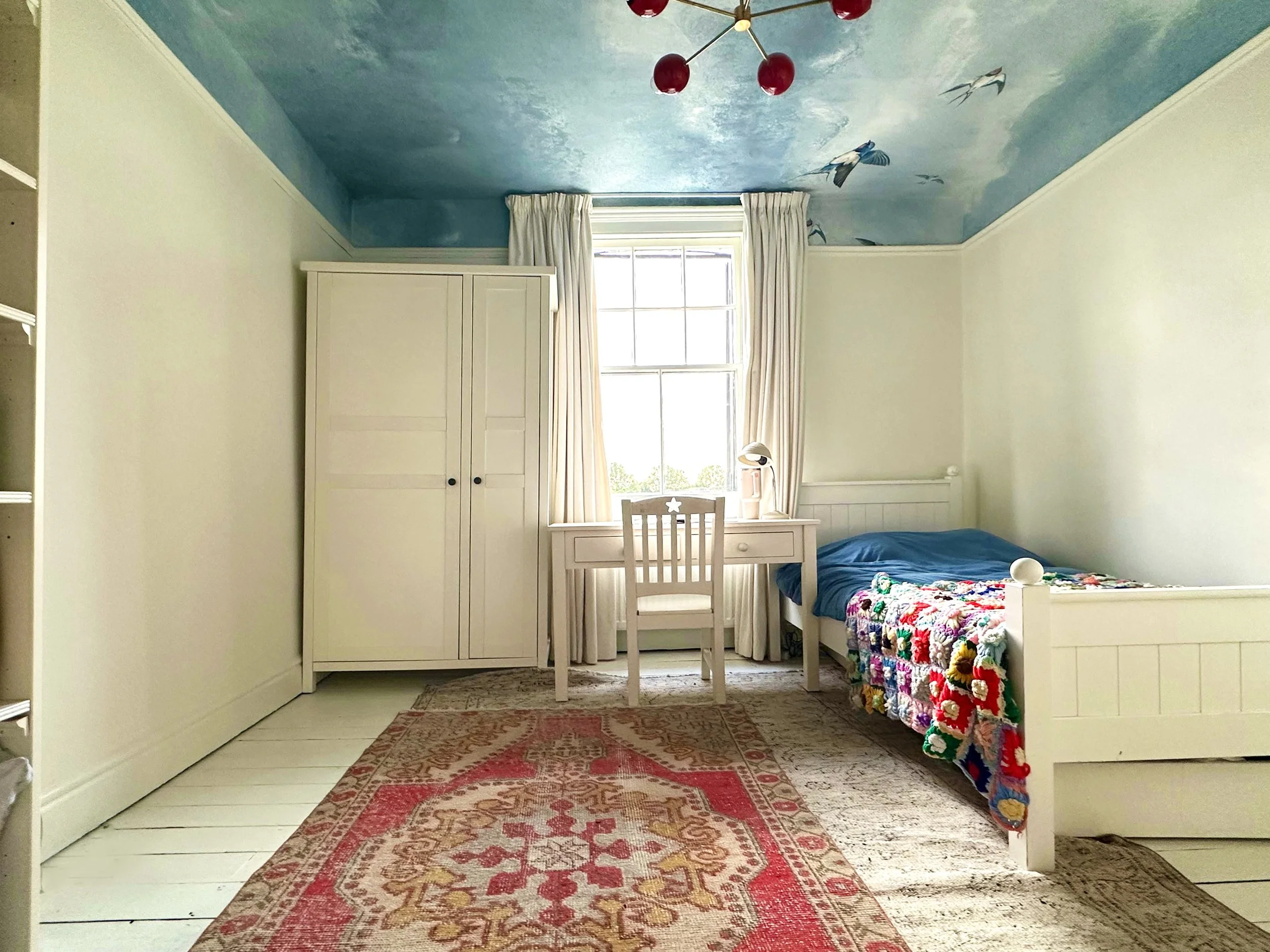 A cozy bedroom featuring a white bed with a colorful crochet blanket, a white desk with a lamp, a white wardrobe, and a window with cream curtains. The ceiling is painted with blue sky and clouds, with painted birds flying.