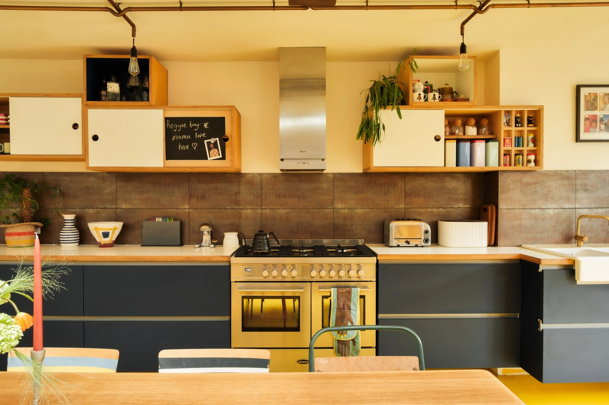A modern kitchen with a yellow oven, black lower cabinets, open shelving holding various kitchen items, a small chalkboard with writing, and a dining table with chairs.