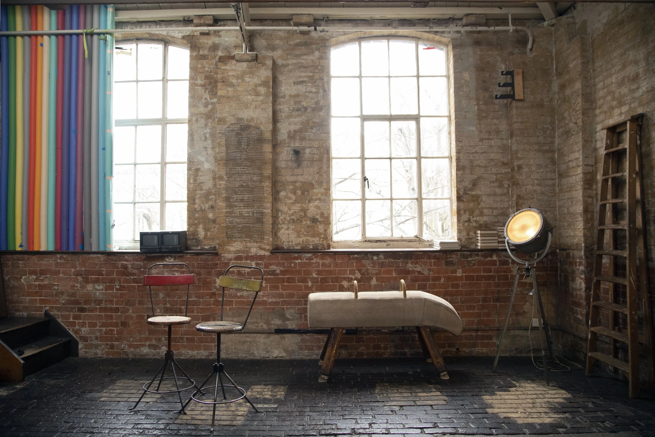 Interior of an industrial-style room with brick walls and large window with natural light, colorful vertical curtains on the left, two vintage chairs with worn paint, a beige padded bench or medical examination table, a professional studio spotlight,
