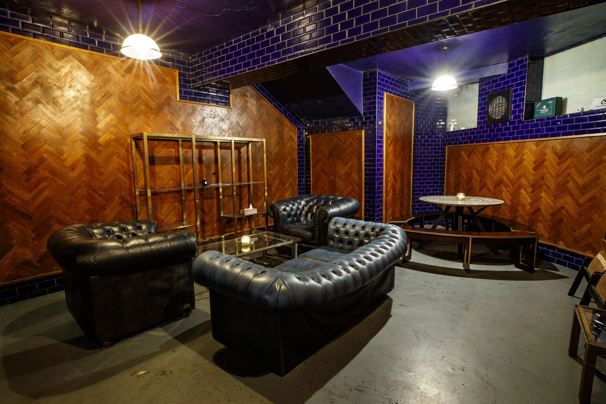  Lounge area with black leather Chesterfield sofas, glass coffee tables, a round marble table, wooden panel walls, and blue tiled accents in the ceiling and walls, illuminated by hanging lights.