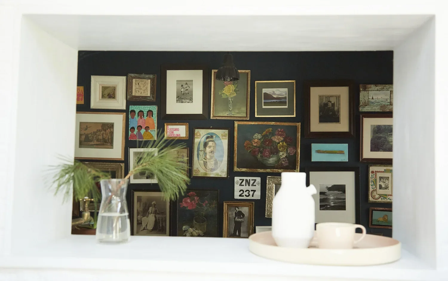 View through a wall opening showing an interior wall decorated with various framed artworks and photos, with a hanging light fixture, and a white shelf with a vase holding greenery, a white pitcher, and a pink cup on a tray in the foreground.