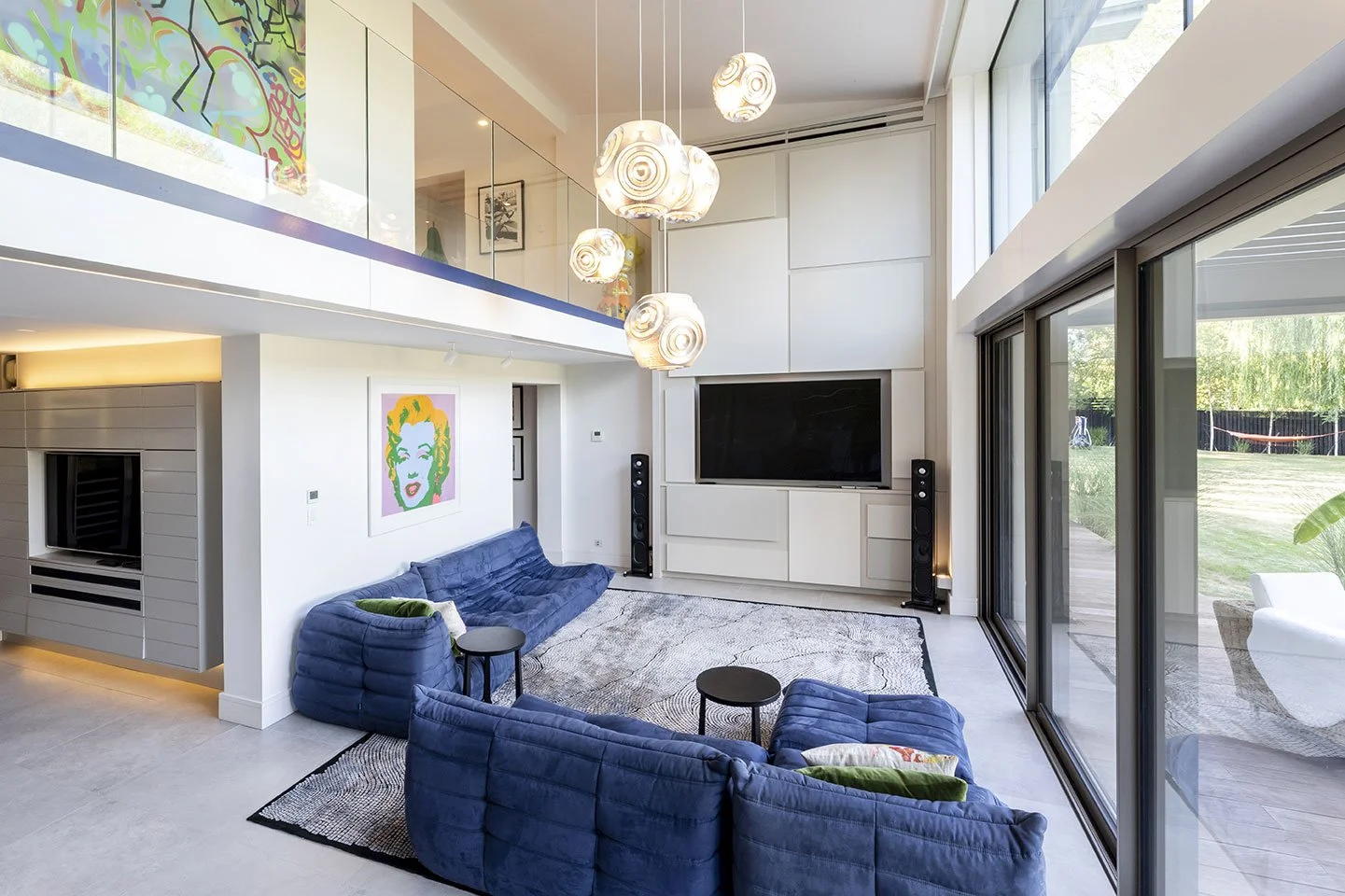 Modern living room with large sliding glass doors, blue couches, a large flat-screen TV, abstract artwork on the wall, and a patterned rug. Ceiling lights hang from above.