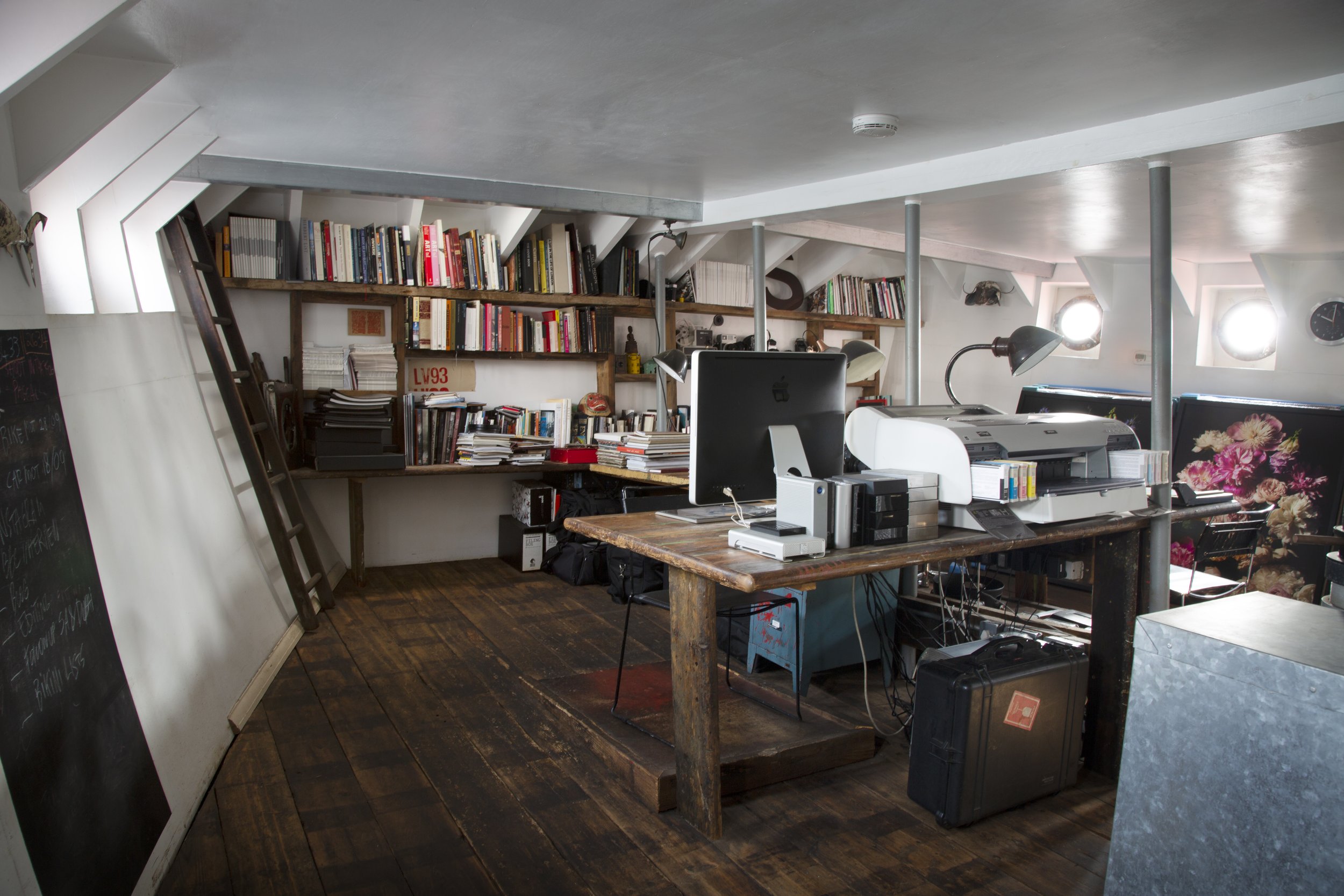 A cozy attic office space with a wooden desk holding a computer, printers, and office supplies. Shelves filled with books and stacks of papers line the back wall. Natural light streams through small round windows, with a blackboard on the left side a