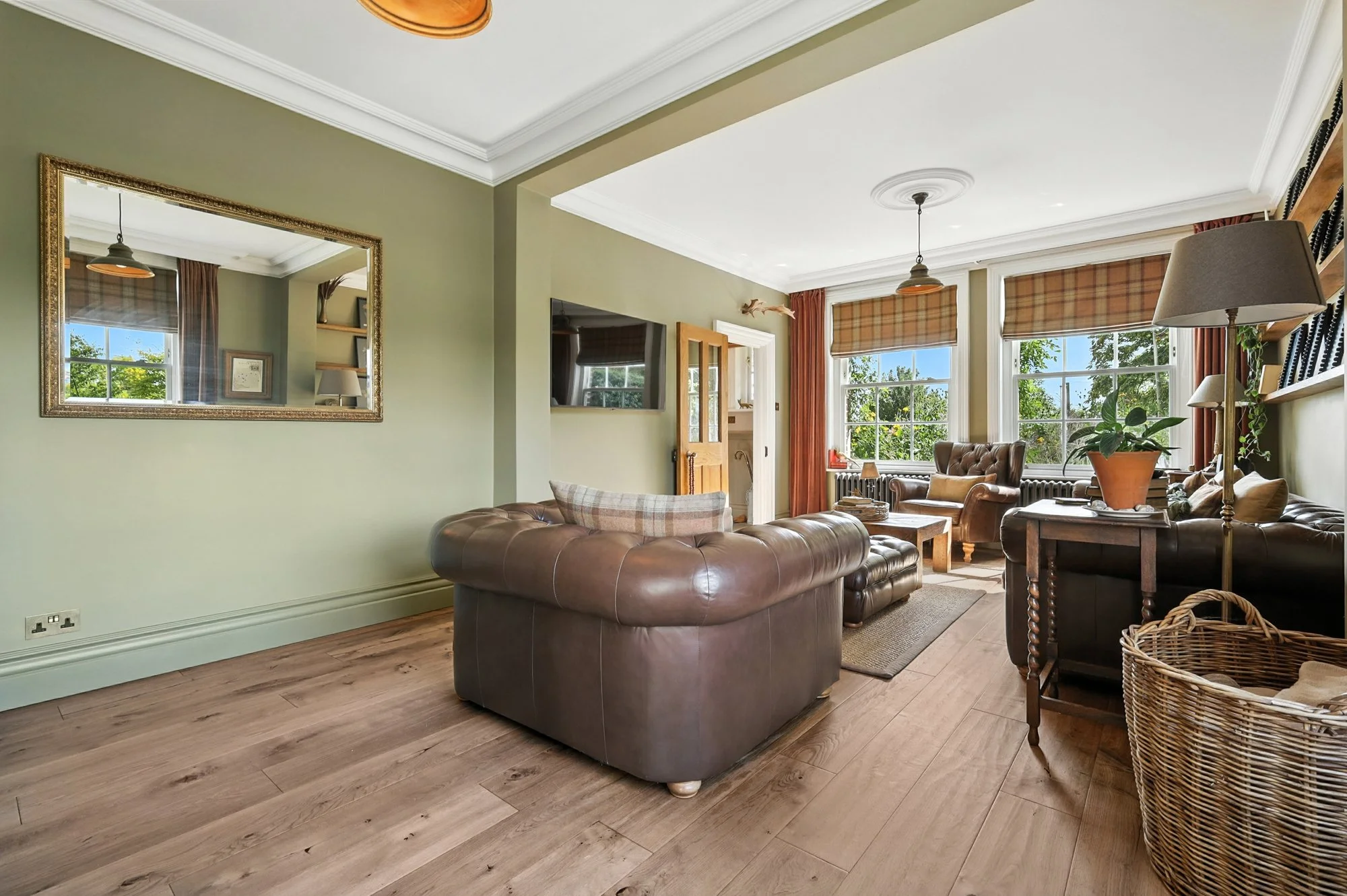 Living room with green walls, hardwood floors, leather sofa, armchair, large windows with plaid curtains, a mirror on the wall, bookshelf, potted plant, and ceiling lights.
