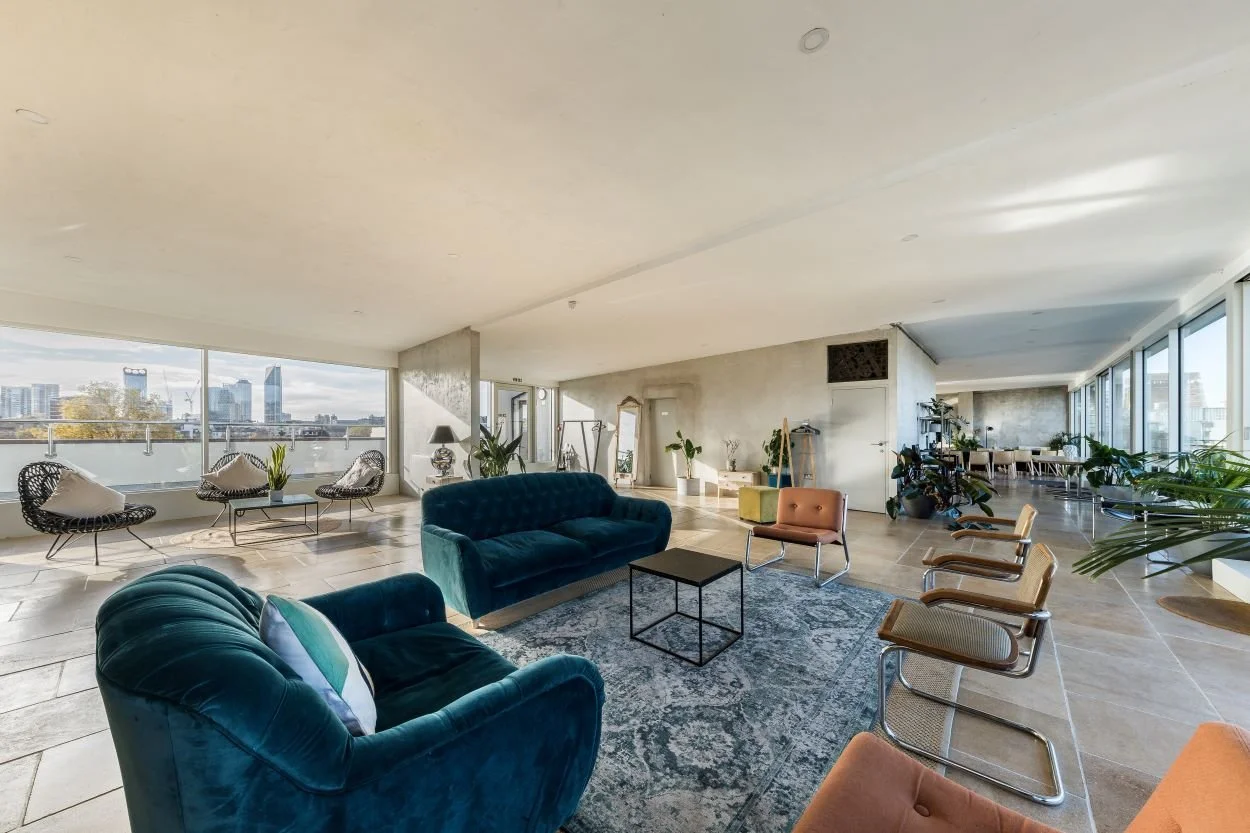 Spacious modern living room with large windows, blue velvet sofas, various chairs, plants, and city skyline view