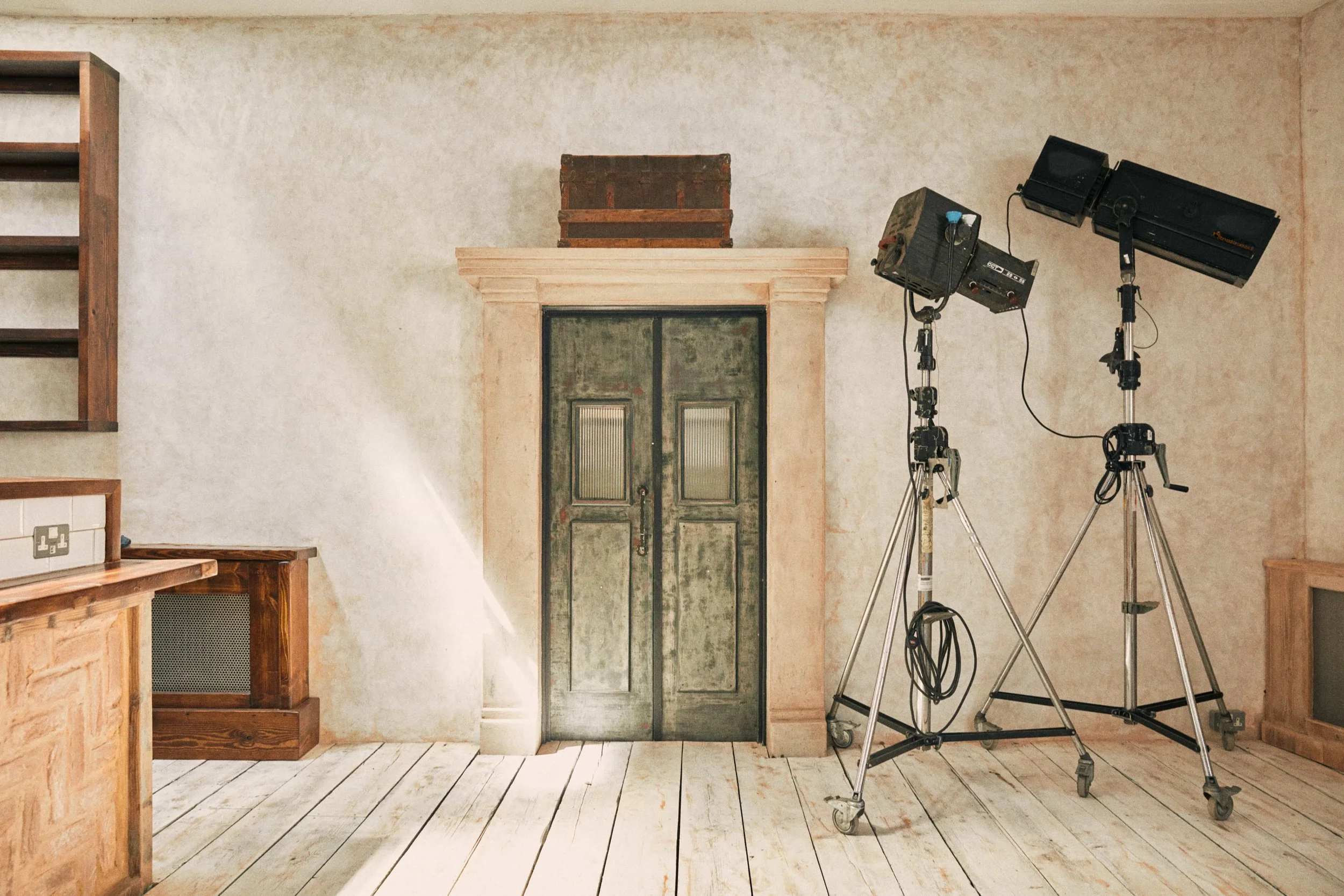 Empty room with studio lighting equipment on tripods, a rustic door, a wooden cabinet, and a wooden staircase. The walls are plain and the floor has white wooden planks.