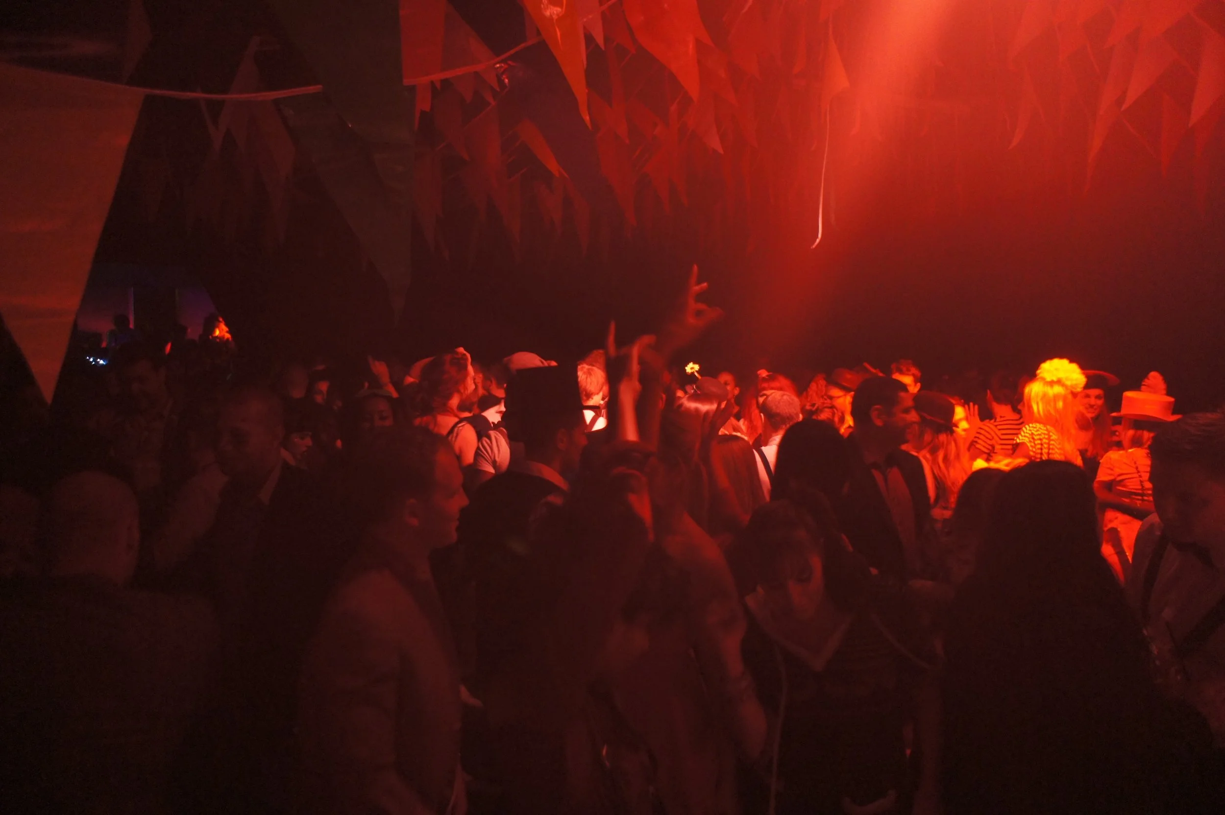 Crowd of people at a concert or club with red lighting and decorative hanging banners.