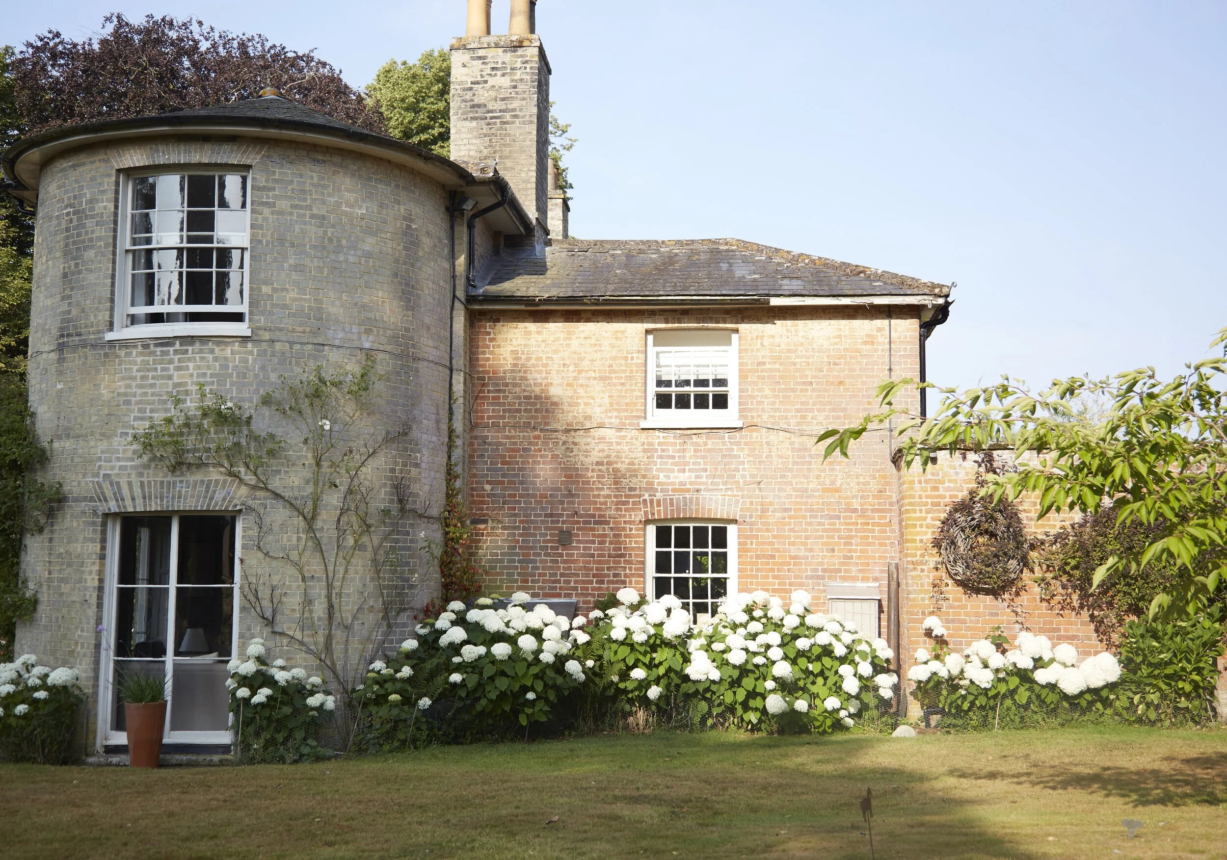 A two-story brick house with a rounded tower section on the left, three white-framed windows, a chimney, and a garden with white hydrangeas in front.