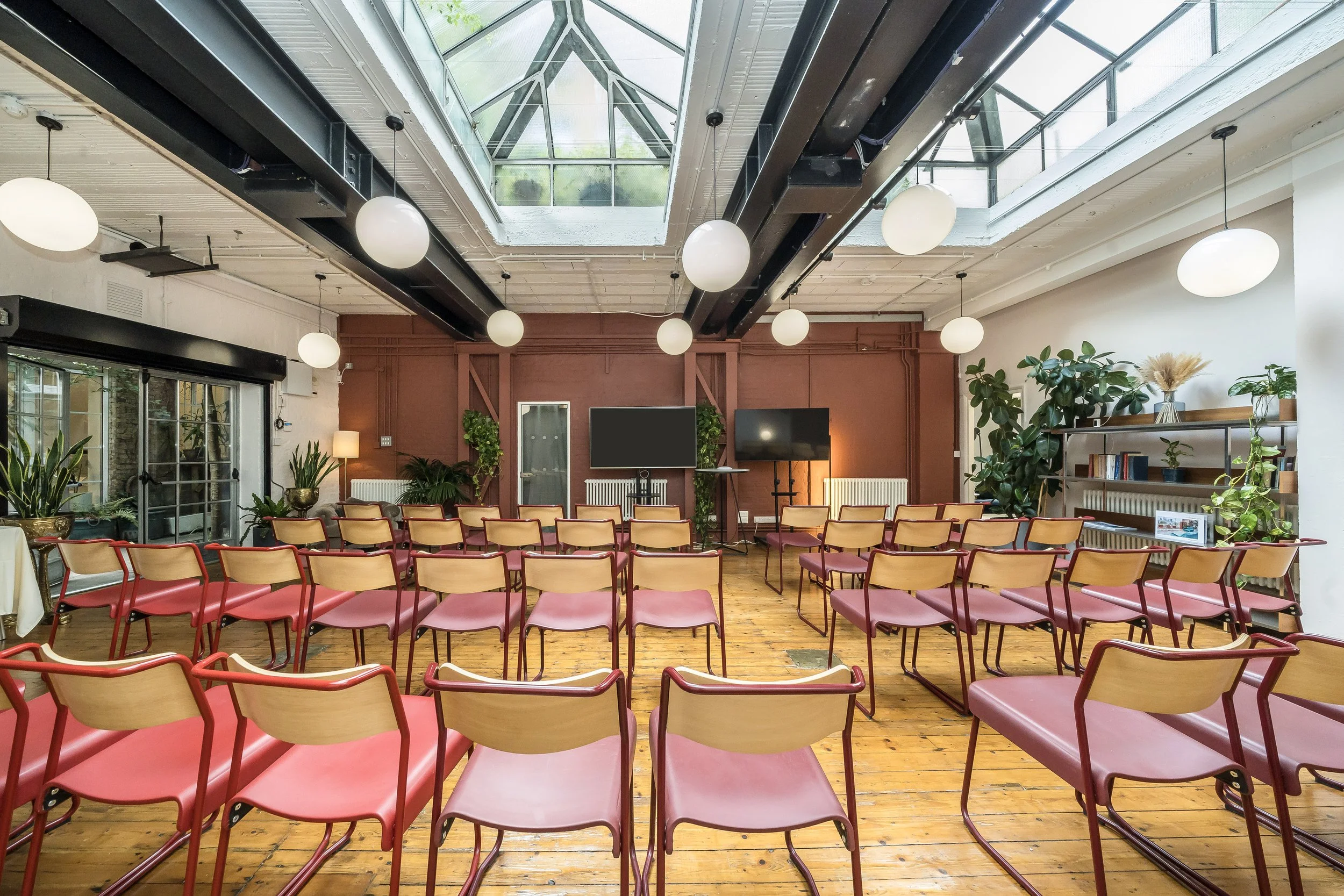 Conference room with rows of red chairs facing two television screens, plants, bookshelves, and large windows with a glass ceiling overhead
