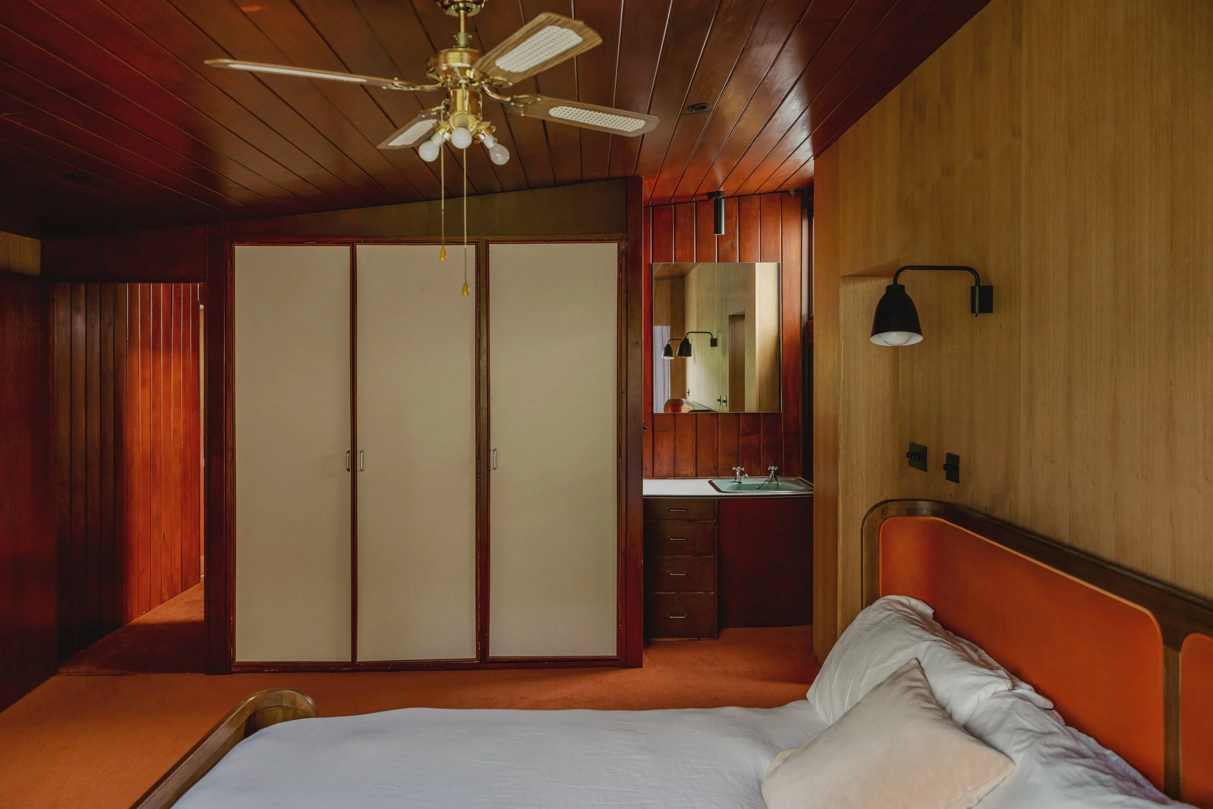 A bedroom with wood-paneled walls and ceiling, a bed with white bedding, a nightstand, and a small kitchenette with a sink and cabinets. A ceiling fan with lights is mounted on the ceiling.