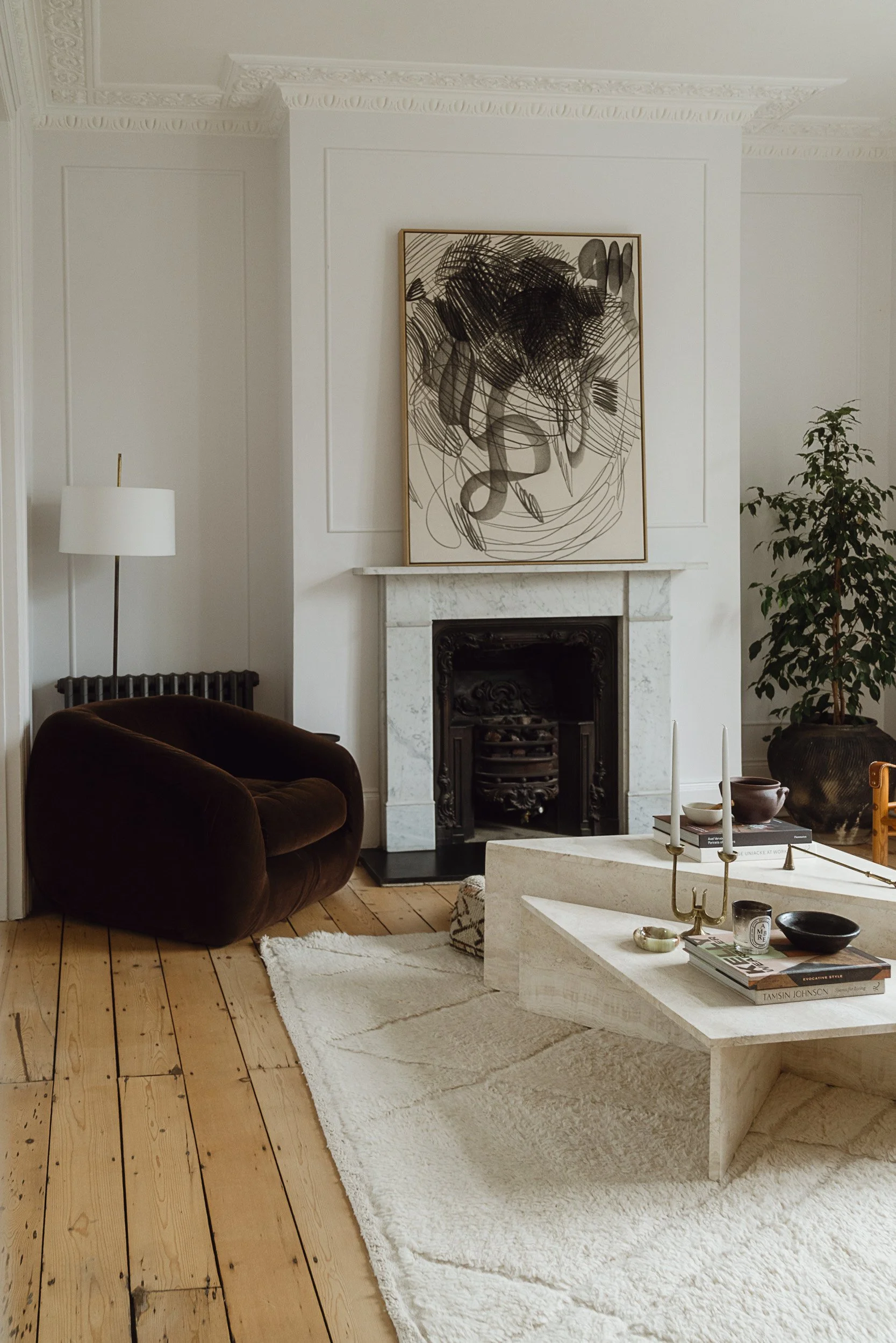 Living room with a white fireplace, abstract black and white artwork above, brown velvet armchair, white textured rug, cream marble coffee table with books, bowls, and candlesticks, and a tall potted plant in the corner.