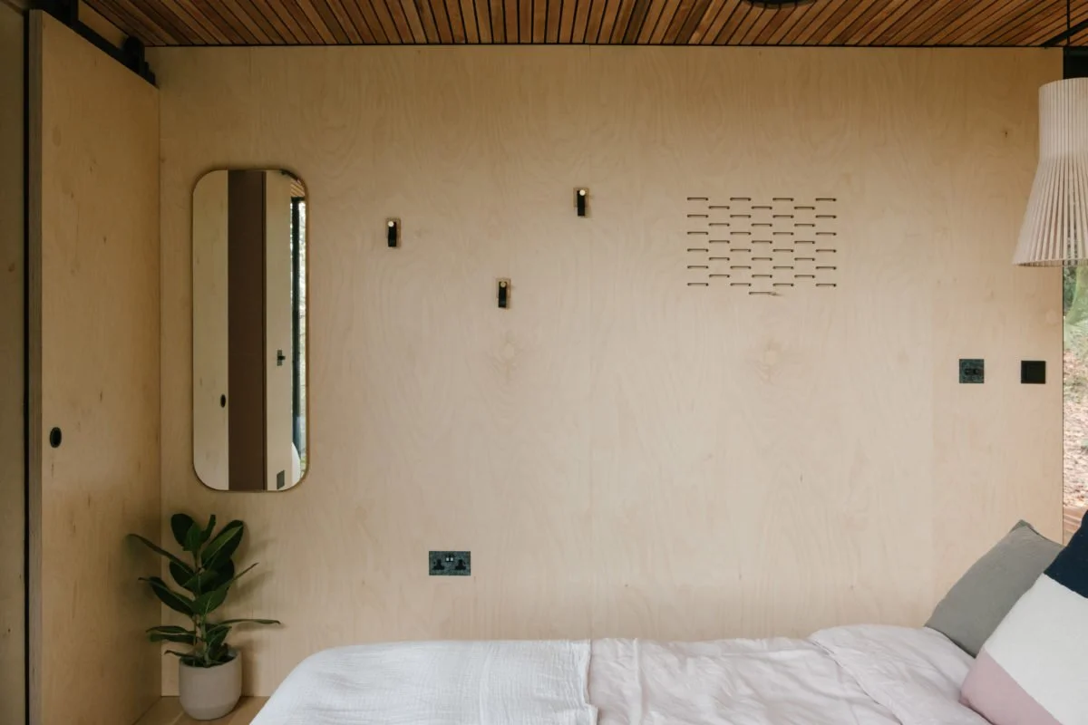 Minimalist bedroom with plain wooden wall, a small mirror, a potted plant, a bed with pillows, and wall fixtures including vent, lights, and switches.