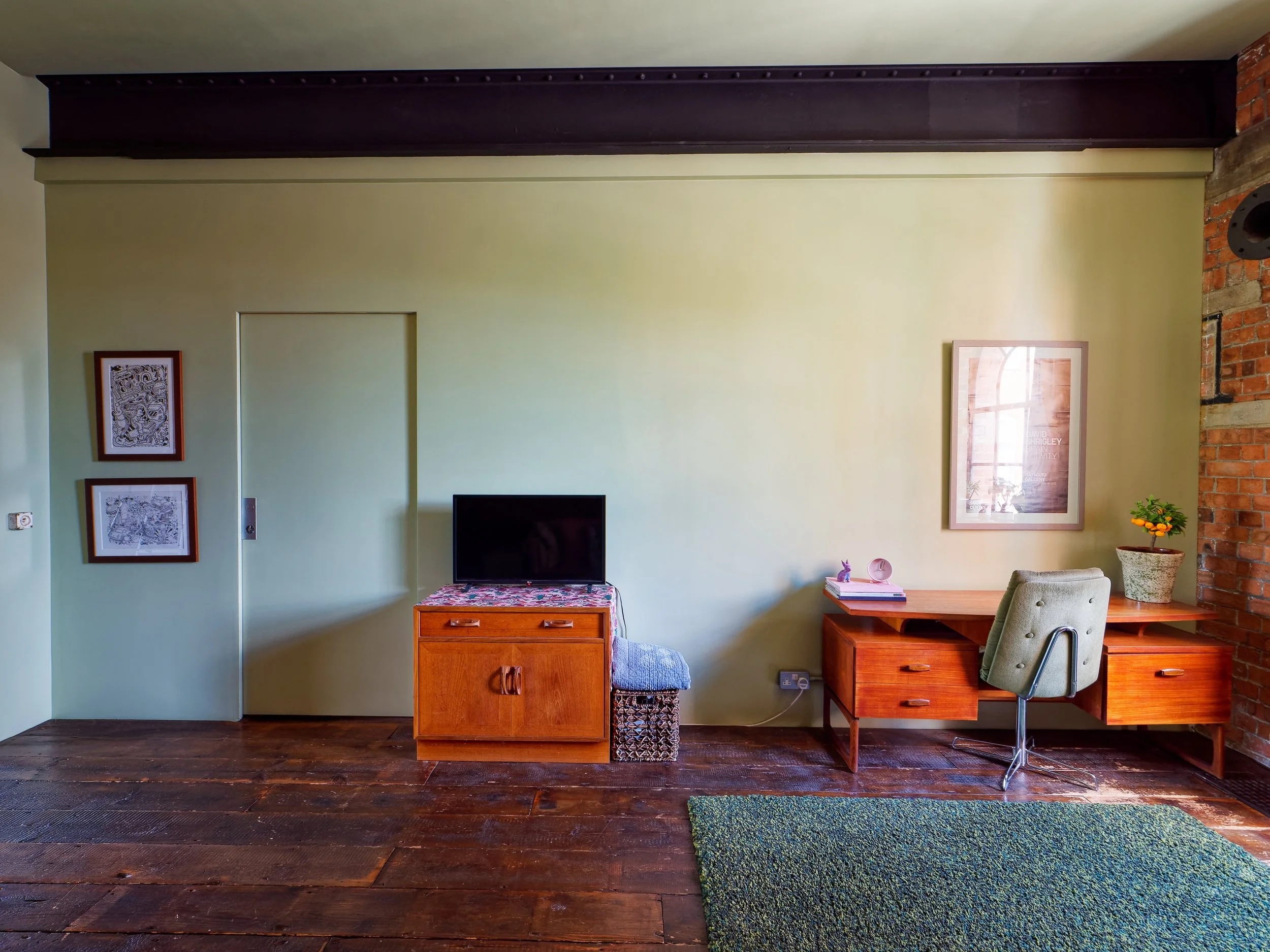 Interior of a room with a green wall, wooden floor, a small television on a wooden cabinet, a desk with a chair, framed pictures, a window reflection, a potted plant, and a green rug.