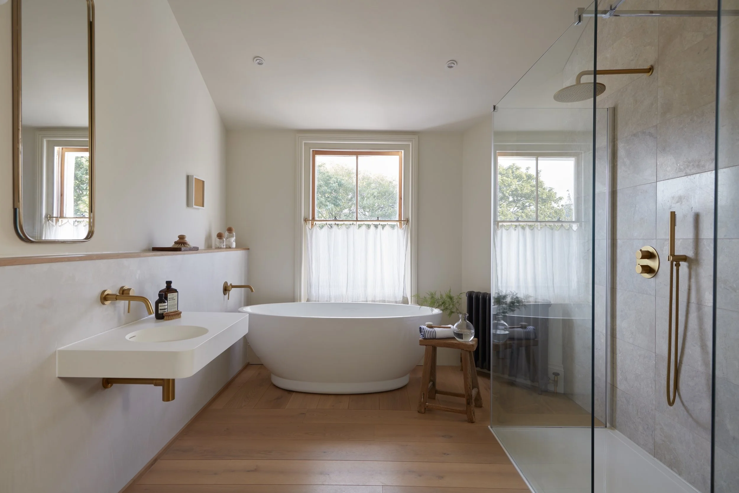 Modern bathroom with free-standing oval bathtub in front of two windows with sheer curtains, a walk-in shower with glass enclosure, and minimalist decor.