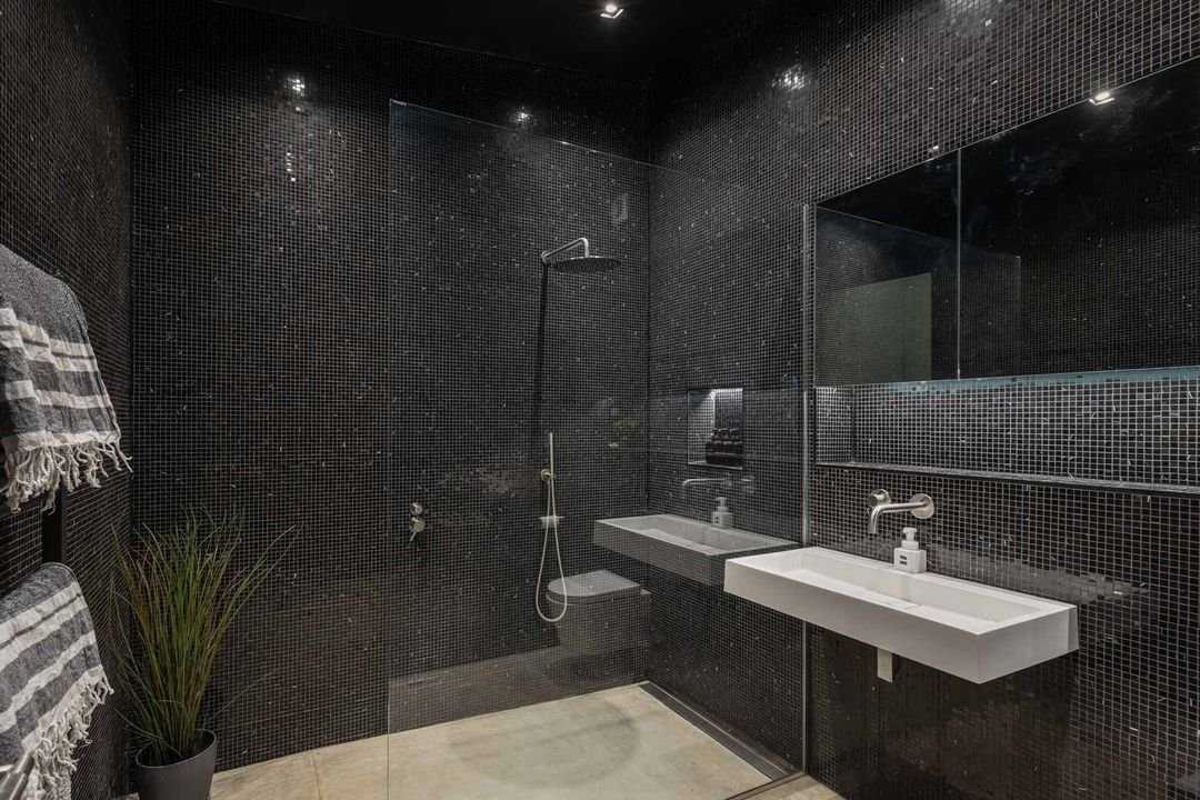 Modern bathroom with black mosaic tile walls, a walk-in shower with rainfall showerhead, a large mirror, two wall-mounted white sinks, a towel rack with striped towels, and a potted plant.
