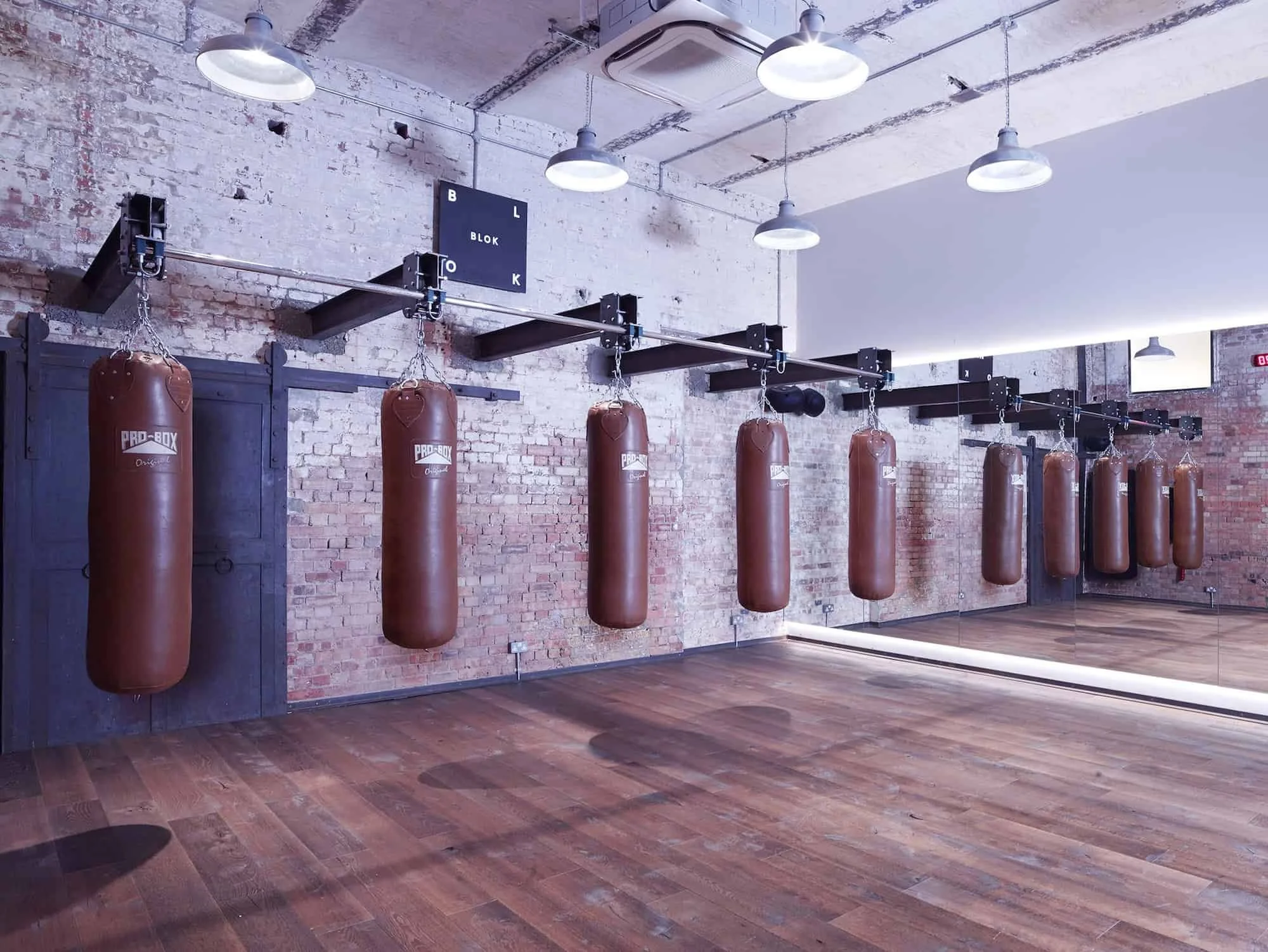 Interior view of a boxing gym with a row of brown punching bags hanging in front of a mirrored wall. The gym has a brick wall, wooden floor, and several ceiling lights.