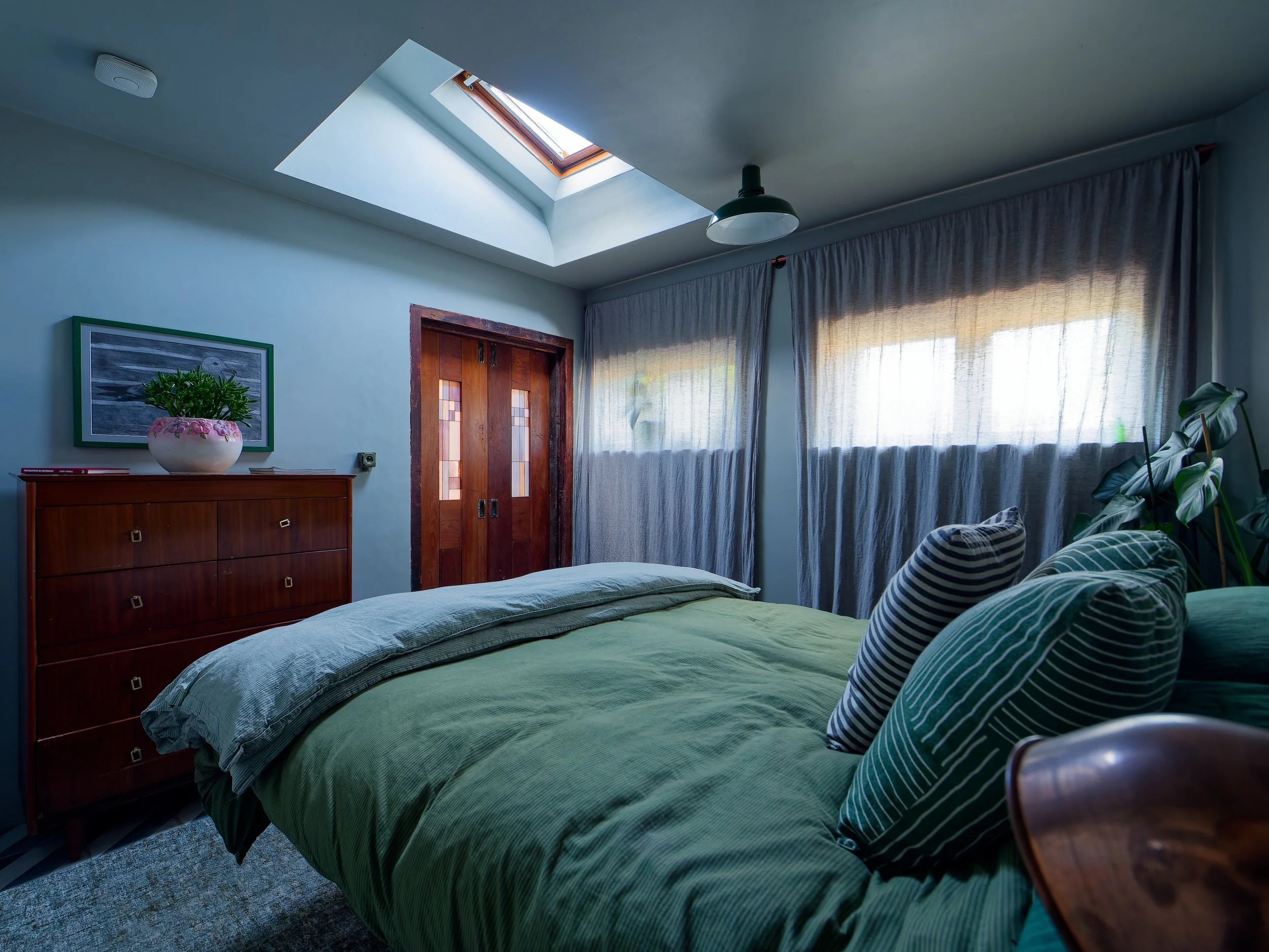 A cozy bedroom with a skylight window, wooden door, dresser with a plant, striped pillows on a bed with green bedding, curtains, and houseplants.