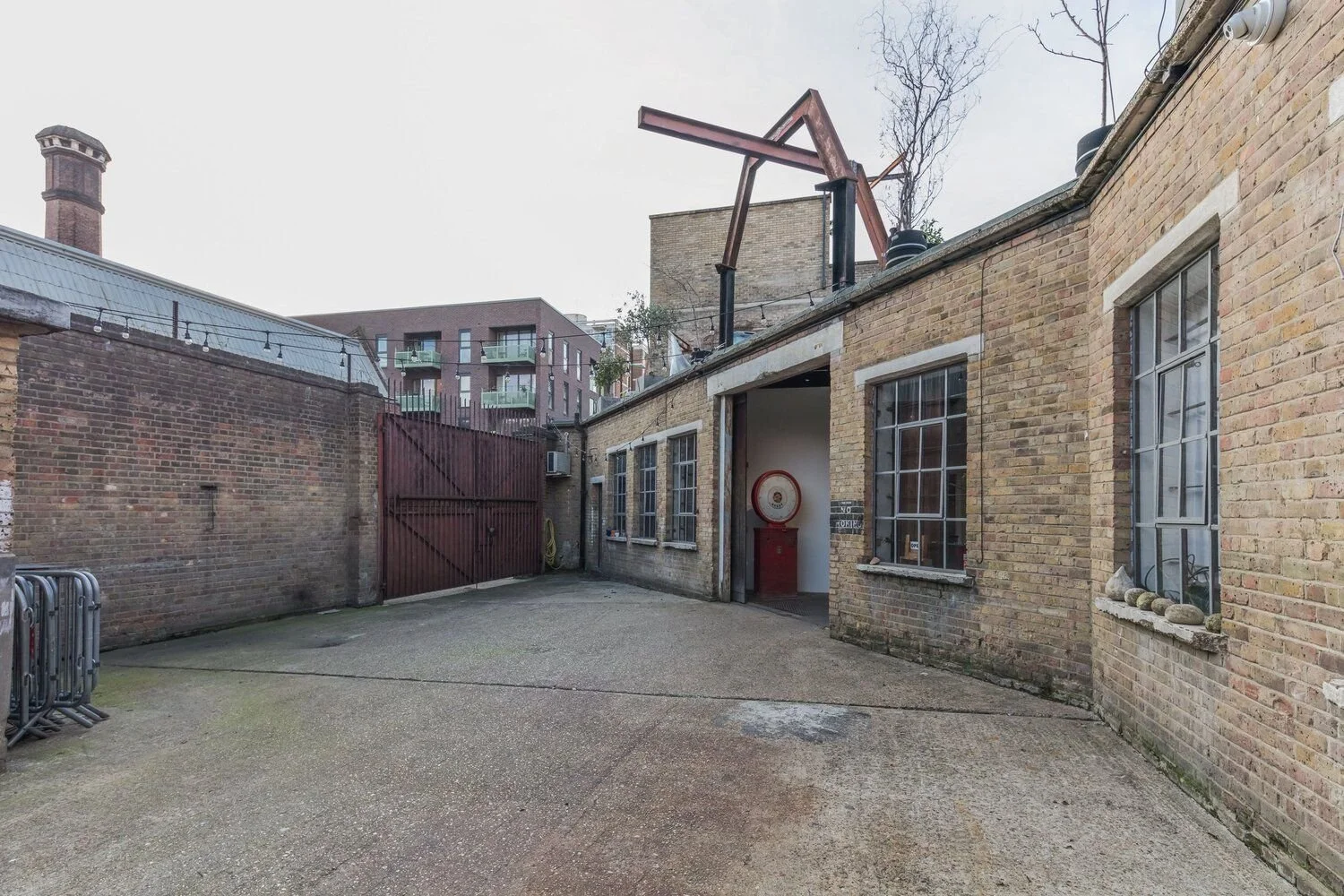 A brick building with multiple windows and a red fire alarm, an open door, and a gate leading to an outdoor courtyard. There's a fire hose reel and some rocks on a window sill, with nearby bicycles and tall modern apartment buildings in the backgroun