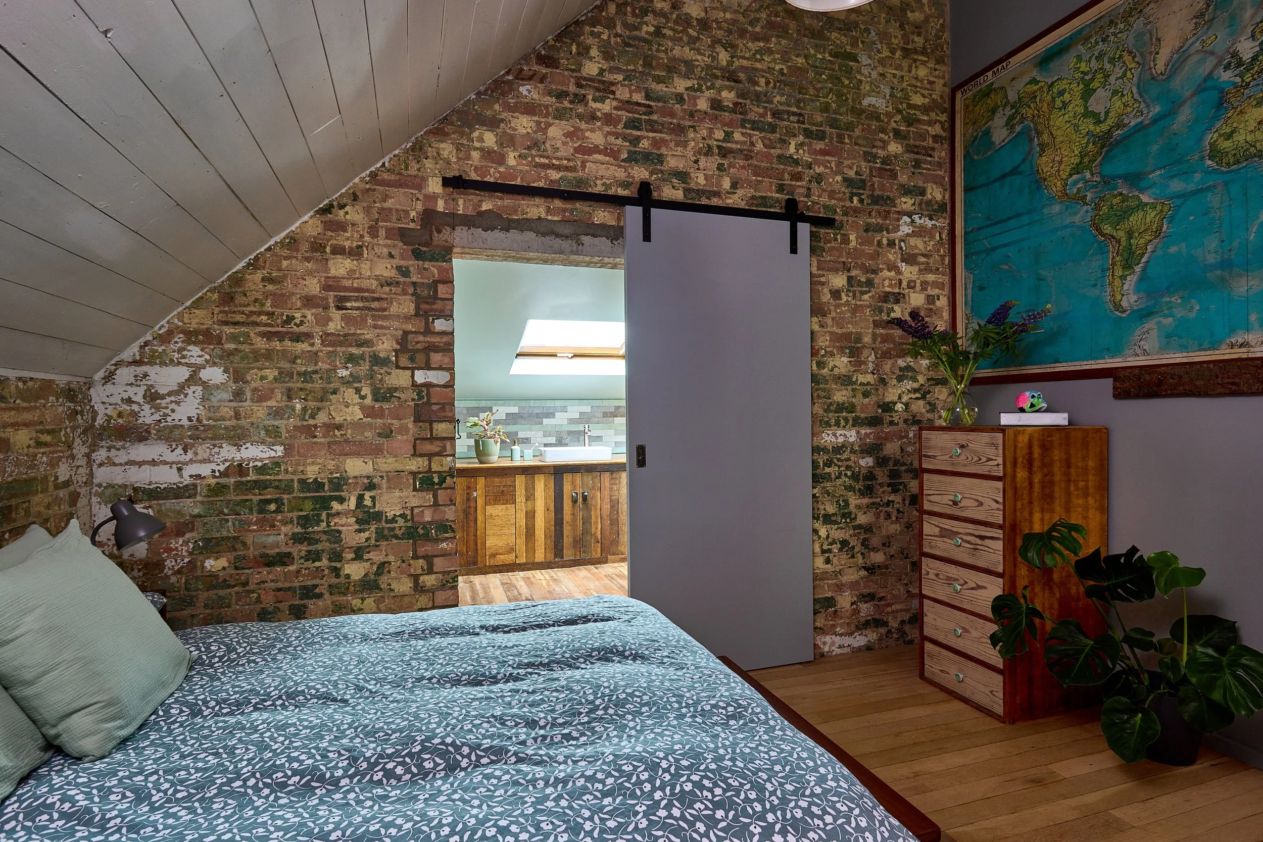 A bedroom with a brick wall, a bed with green pillows, and a sliding barn door leads to a bathroom with a wooden cabinet and skylight. There is a map of the world on the wall and a dresser with plants and a small toy.