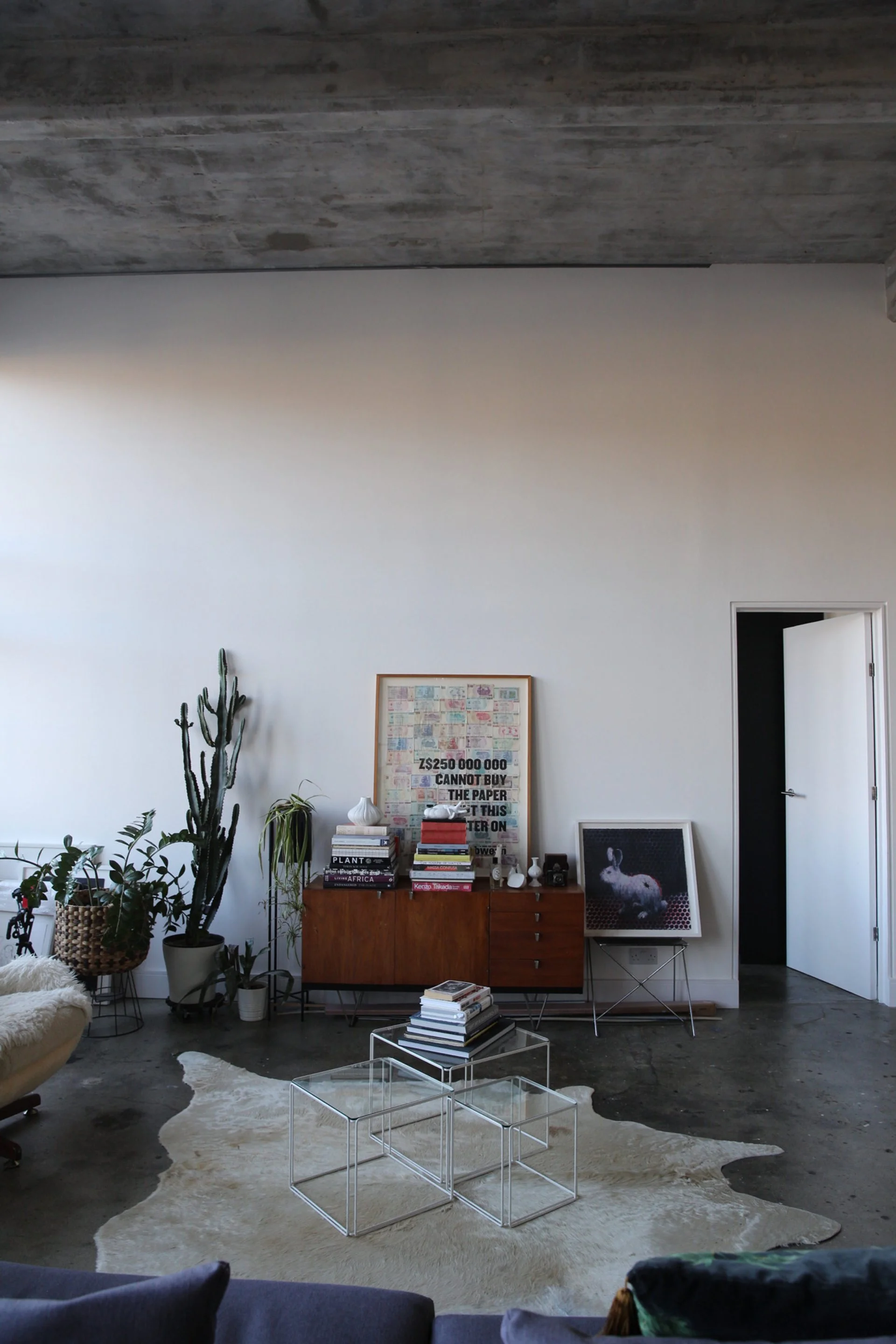 Interior of a modern living room with a white wall, wooden sideboard, and houseplants, a cowhide rug, and contemporary furniture including glass nesting tables and a plush chair.