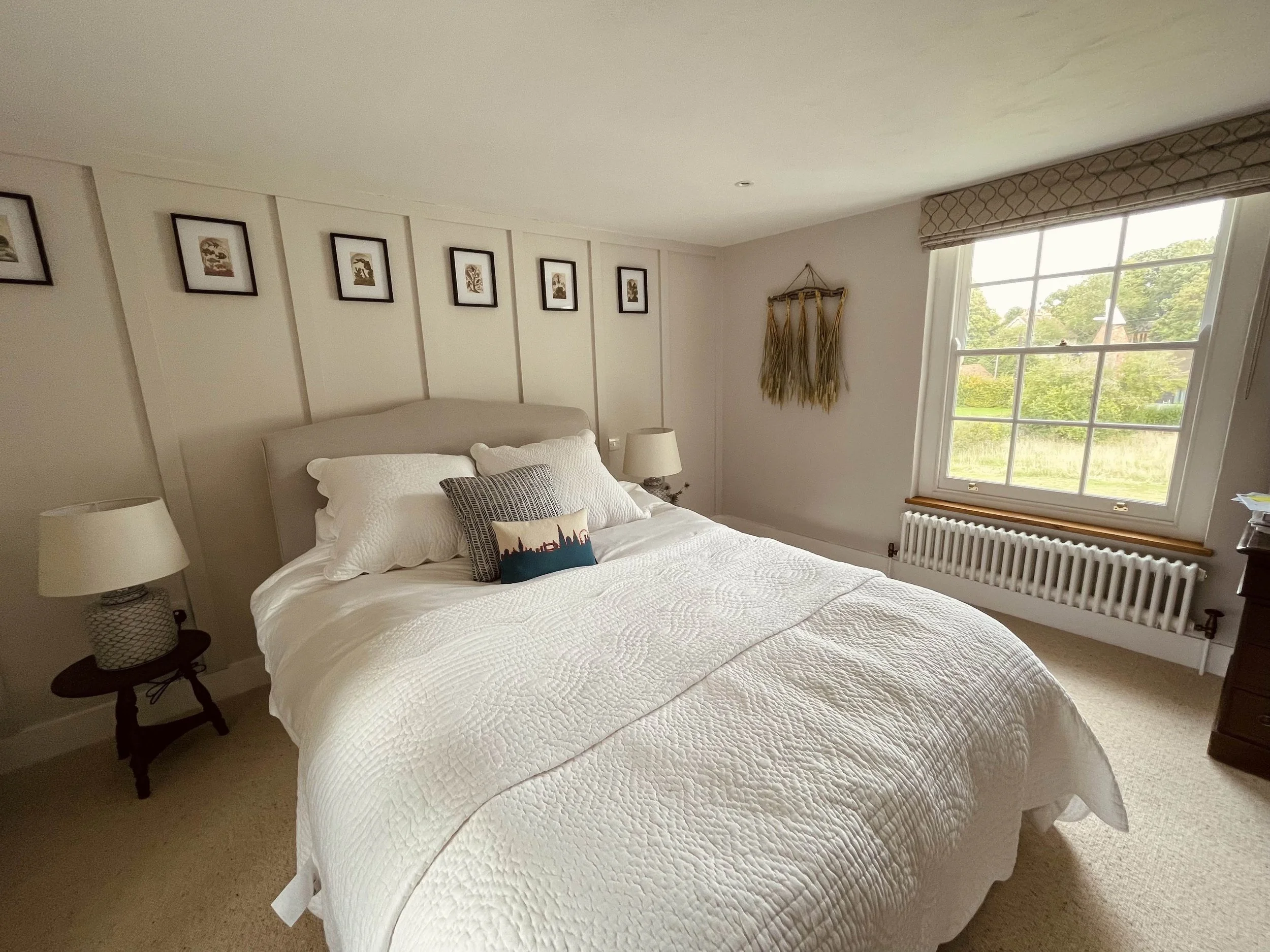 A neatly made bedroom with a large window, white bedding, decorative pillows, and framed artwork on the wall.