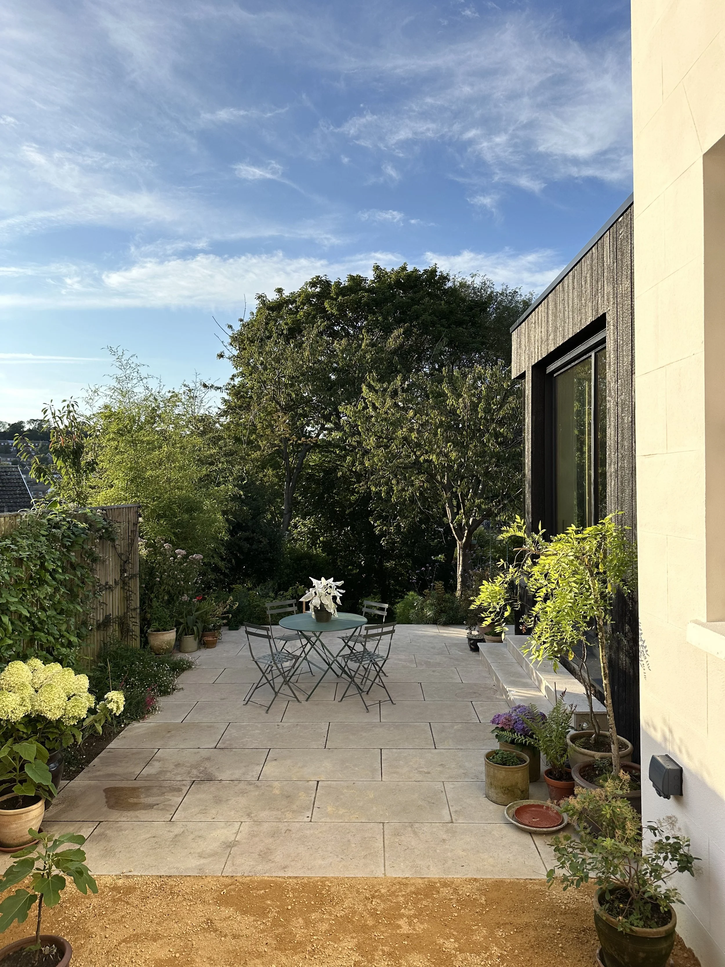 Patio area with potted plants, a small round table with chairs, and trees in the background under a blue sky with scattered clouds.