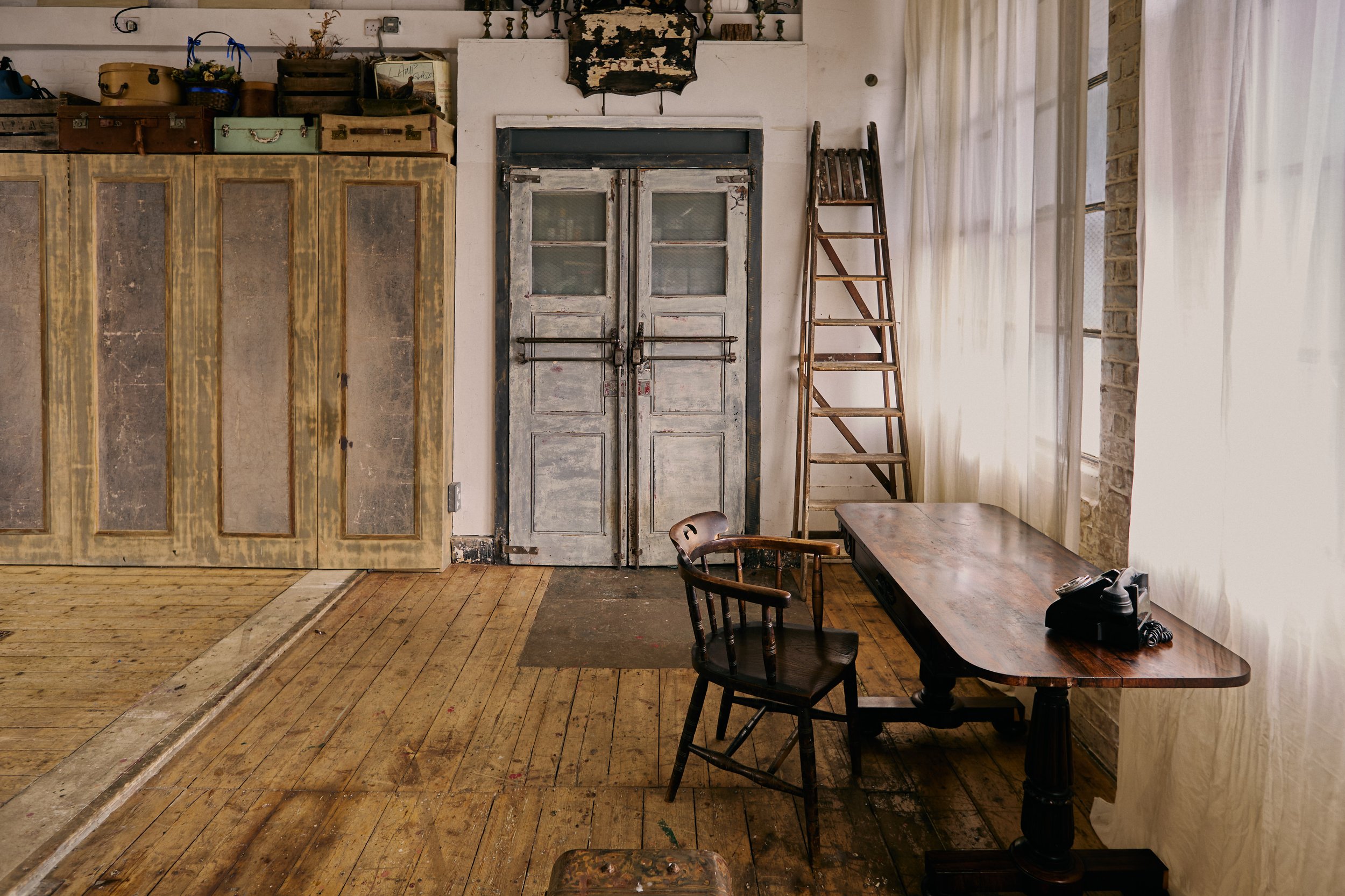Vintage room with wooden floors, old cabinet, closed double doors with chipped paint, ladder, wooden table, and chair, with sunlight filtering through sheer curtains.