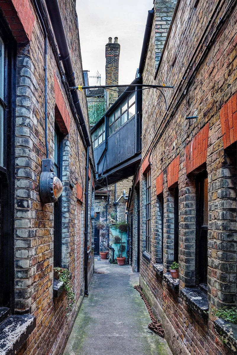 Narrow alleyway between brick buildings with windows, potted plants, and a water fountain at the end, with a chimney visible in the background.