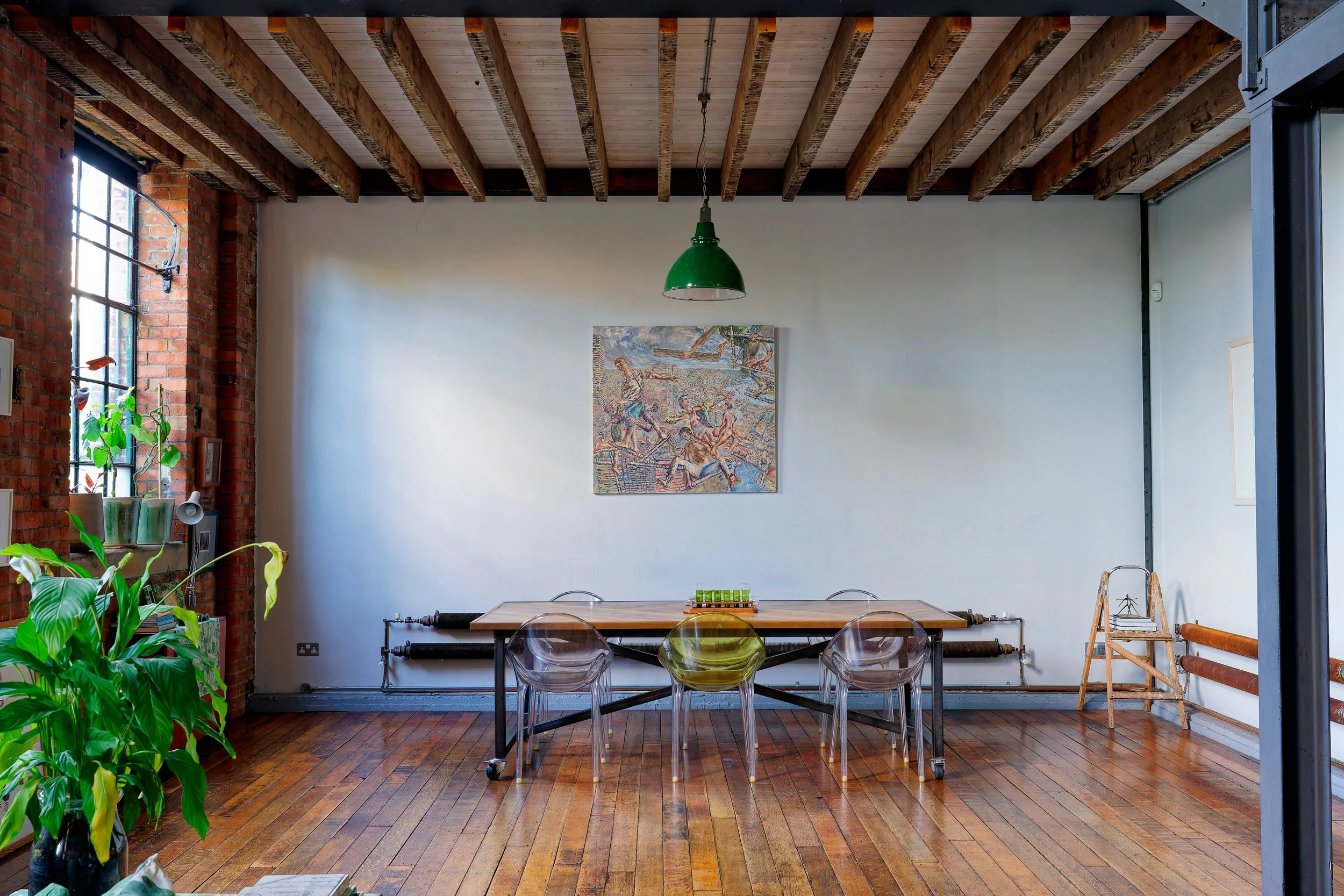 A spacious loft-style room with wooden floors and exposed brick walls. There is a large window on the left side with sunlight coming through, and a green plant on the windowsill. In the center, there's a wooden table with five transparent chairs, one