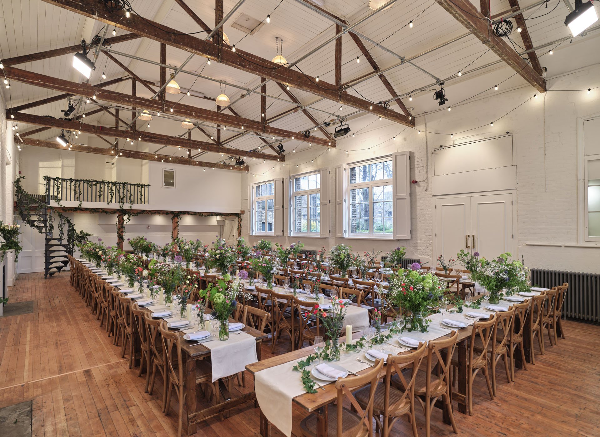 A decorated indoor event space with long wooden tables set for a celebration, adorned with floral centerpieces and white tablecloths, natural light coming through large windows, exposed wooden beams overhead, string lights, and a staircase at the bac