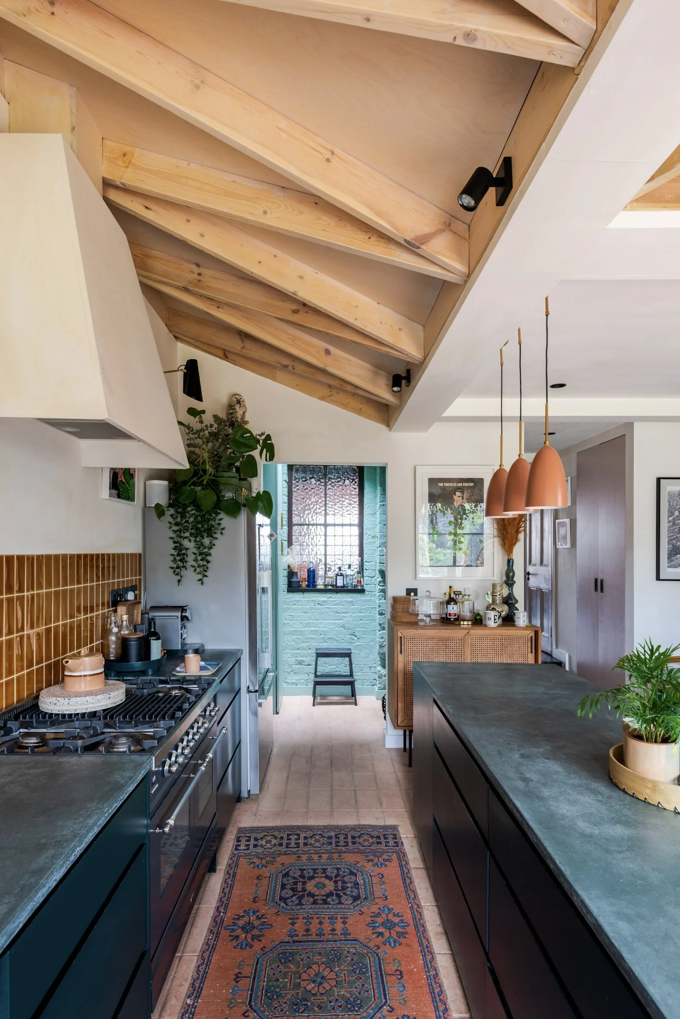 Interior view of a modern kitchen with black cabinets, a green countertop, a rug on the tiled floor, and wooden ceiling beams. Potted plants, hanging pendant lights, and kitchen appliances are visible.