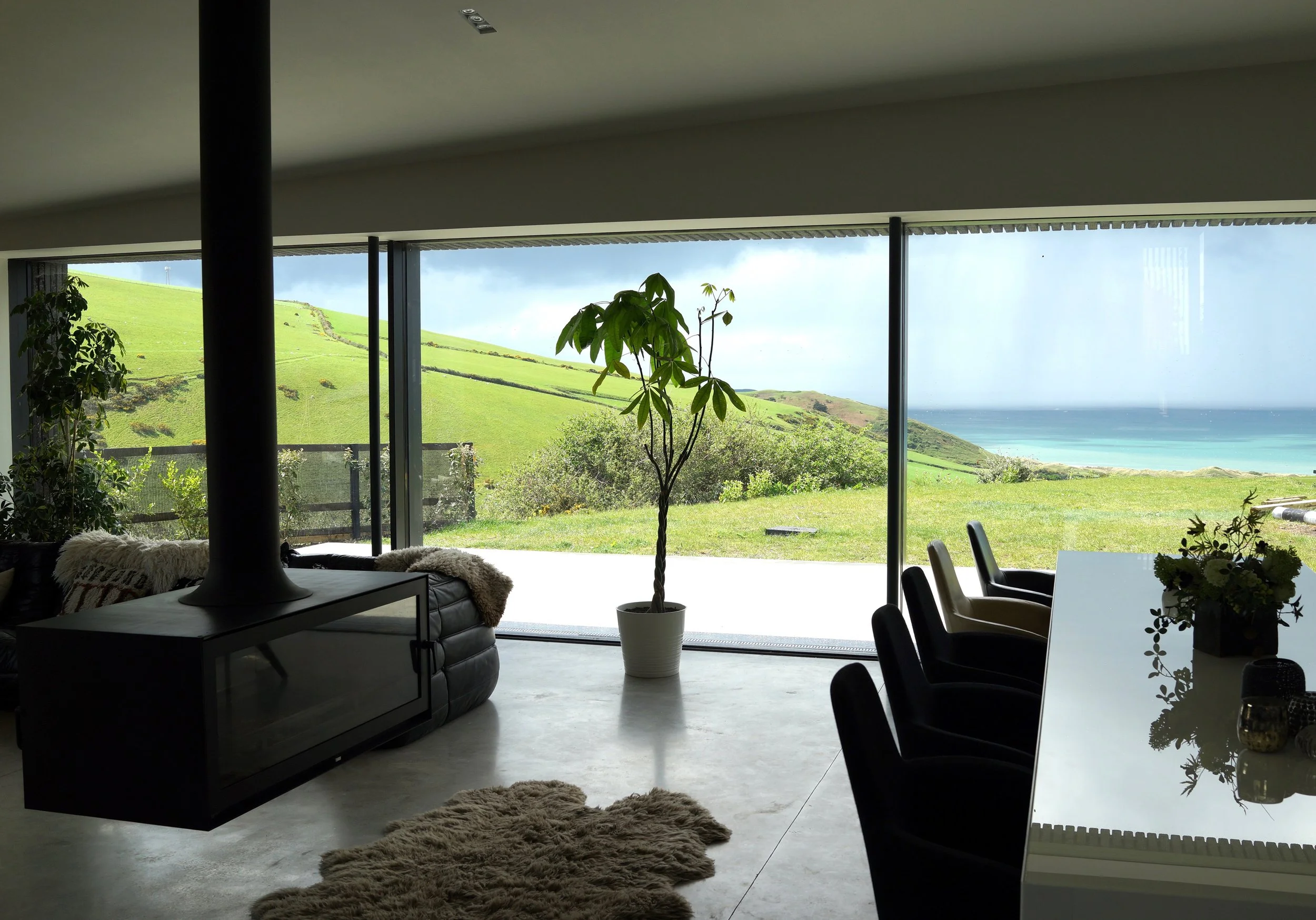 Modern living room with large glass window overlooking green hills and ocean, with a tall potted plant, black chairs, a white table, and cozy decor.