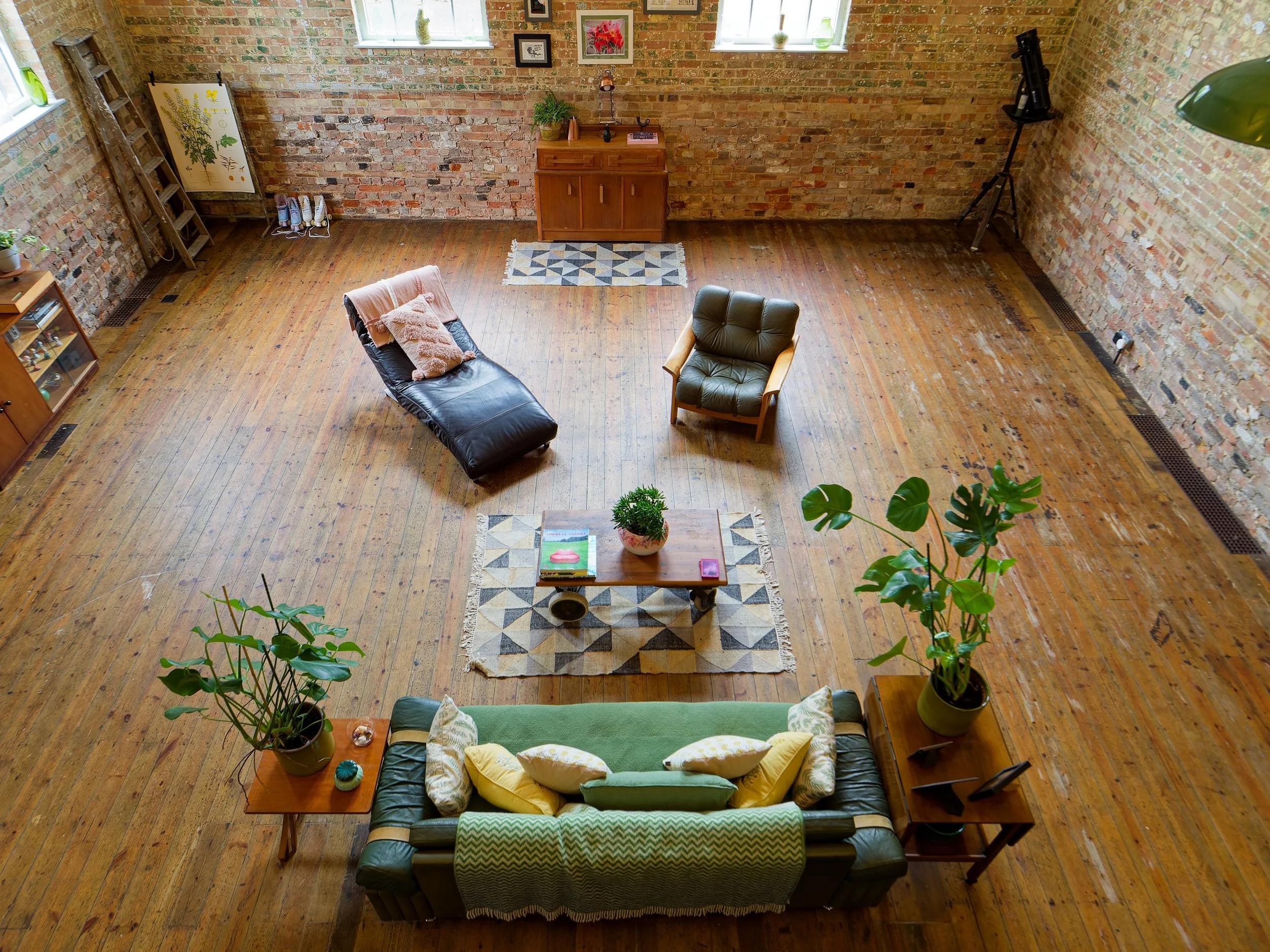 An aerial view of a living room with wooden floors and brick walls. The room has a green sofa with yellow and green pillows, a leather armchair, and a black leather chaise lounge. There are two wooden side tables with plants and other items. In the c