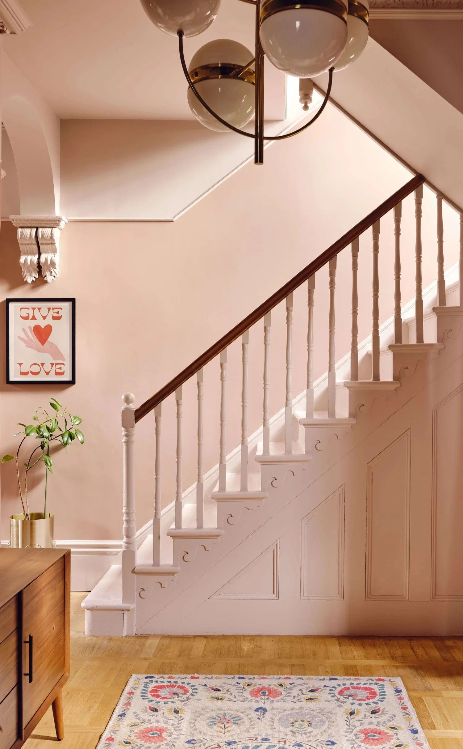 Interior of a home with a staircase, a framed artwork with the word 'LOVE', a potted plant, a wooden console table, a colorful rug, and a ceiling light fixture.