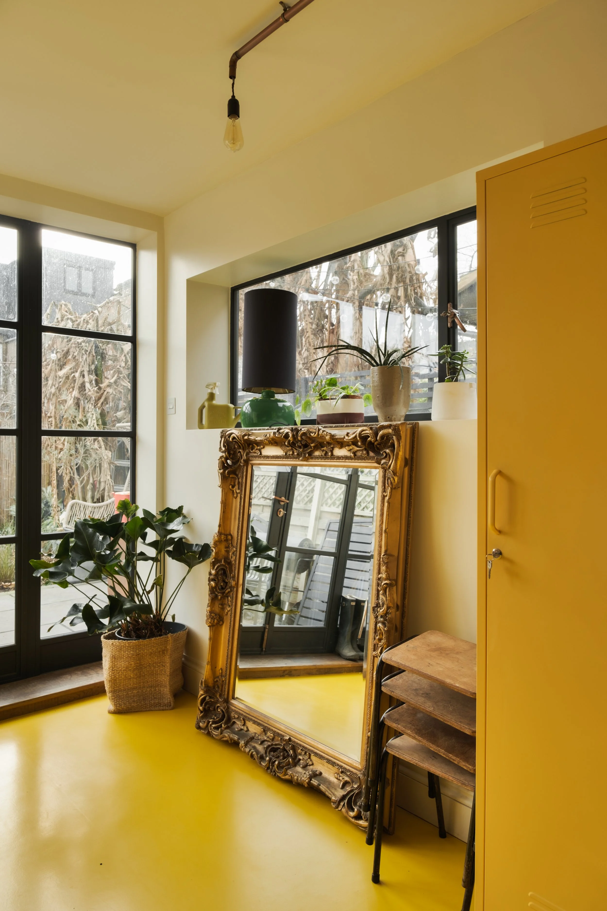 Sunlit room with yellow floors, large black-framed windows, plants on windowsill, gilded mirror leaning against wall, yellow storage cabinet, and small wooden table or stool.