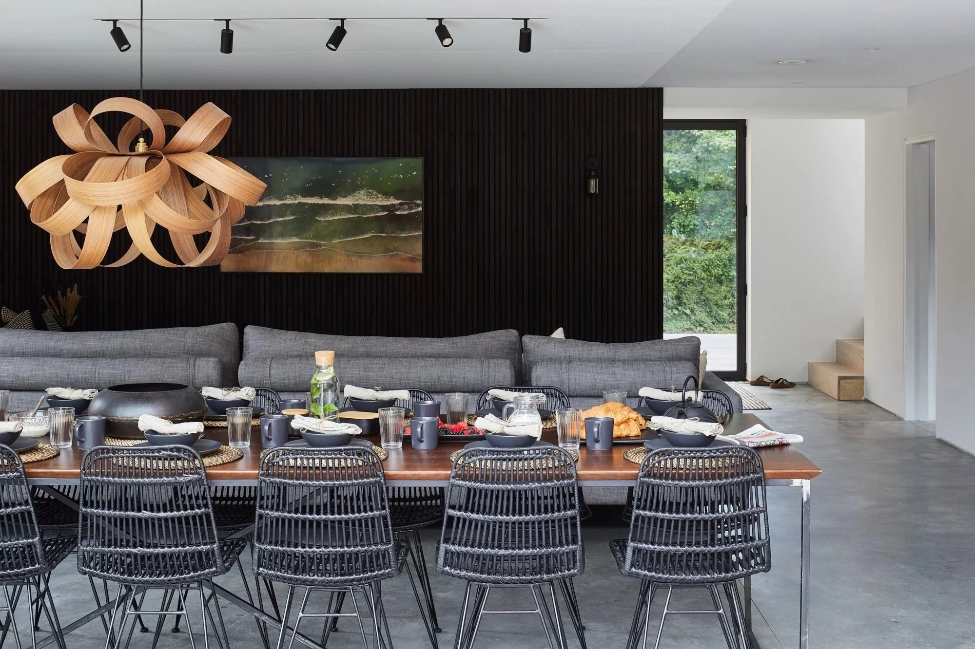 Modern dining room with a large wooden table set with black bowls, cups, and glasses, surrounded by black wire chairs, gray sectional sofa, black wooden paneled wall, abstract painting, and unique wooden chandelier.