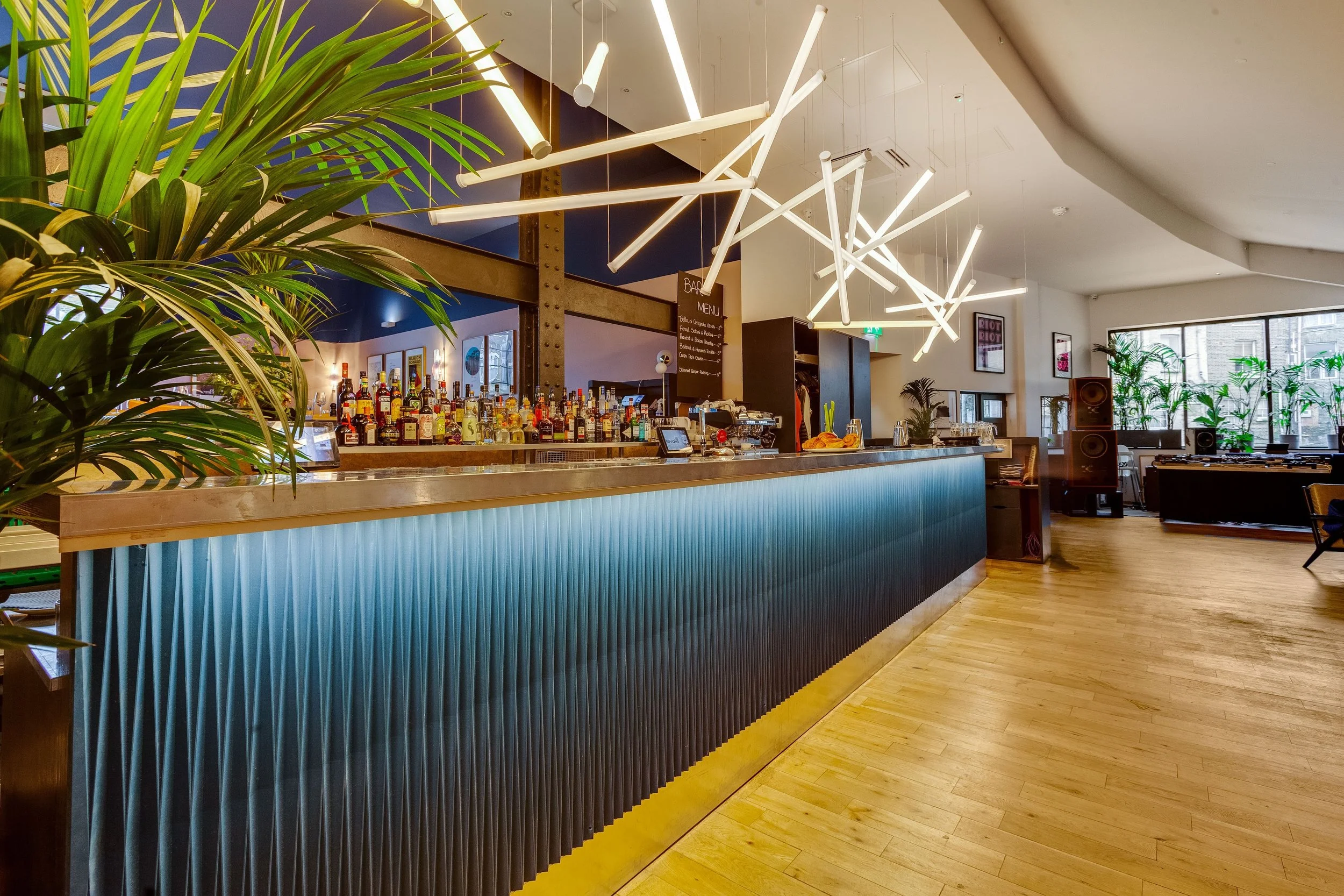 Modern bar with illuminated textured front, bottles on shelves, abstract white ceiling lights, large window with plants, and wooden flooring.