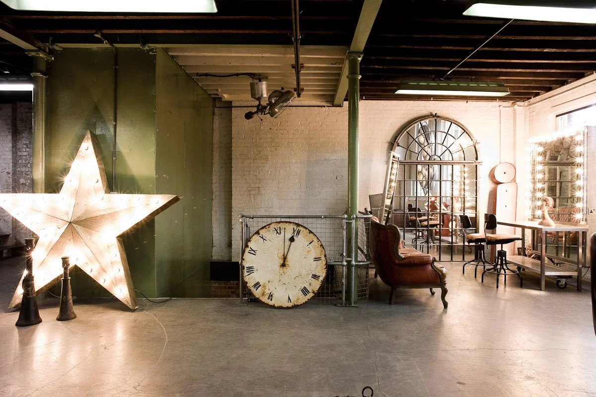 Interior of a decorated room featuring a large illuminated star on the left, vintage-style clock, a red armchair, a large arched window, and a vanity mirror with lights on the right.