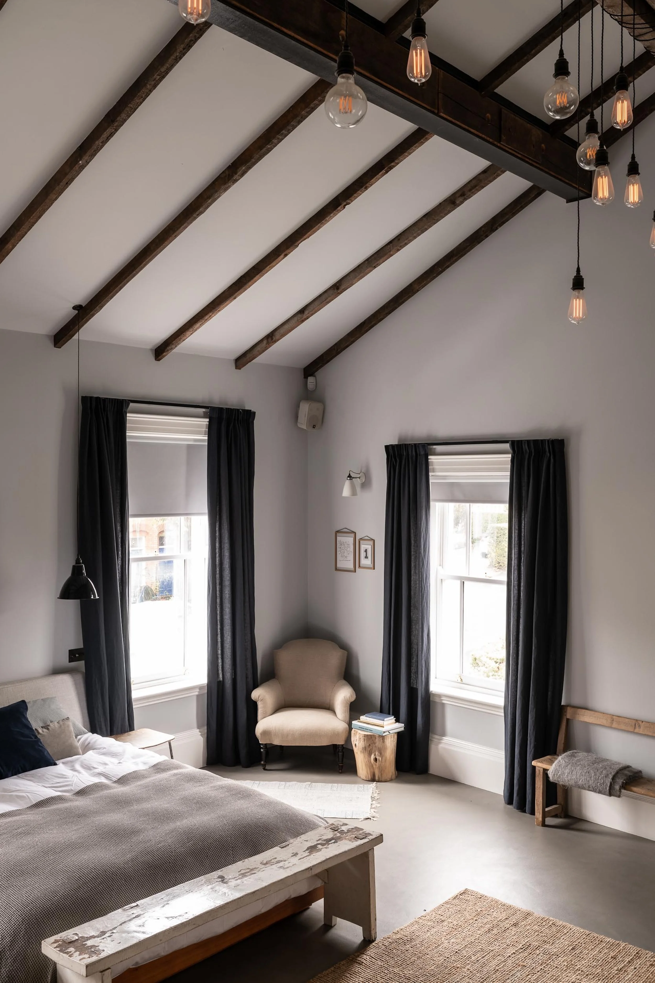 A bedroom with a high, white ceiling and dark wooden beams. There are hanging Edison bulb lights, a beige armchair near two windows with dark curtains, a bed with a light-colored headboard, and a small wooden bench at the foot of the bed. A side tabl